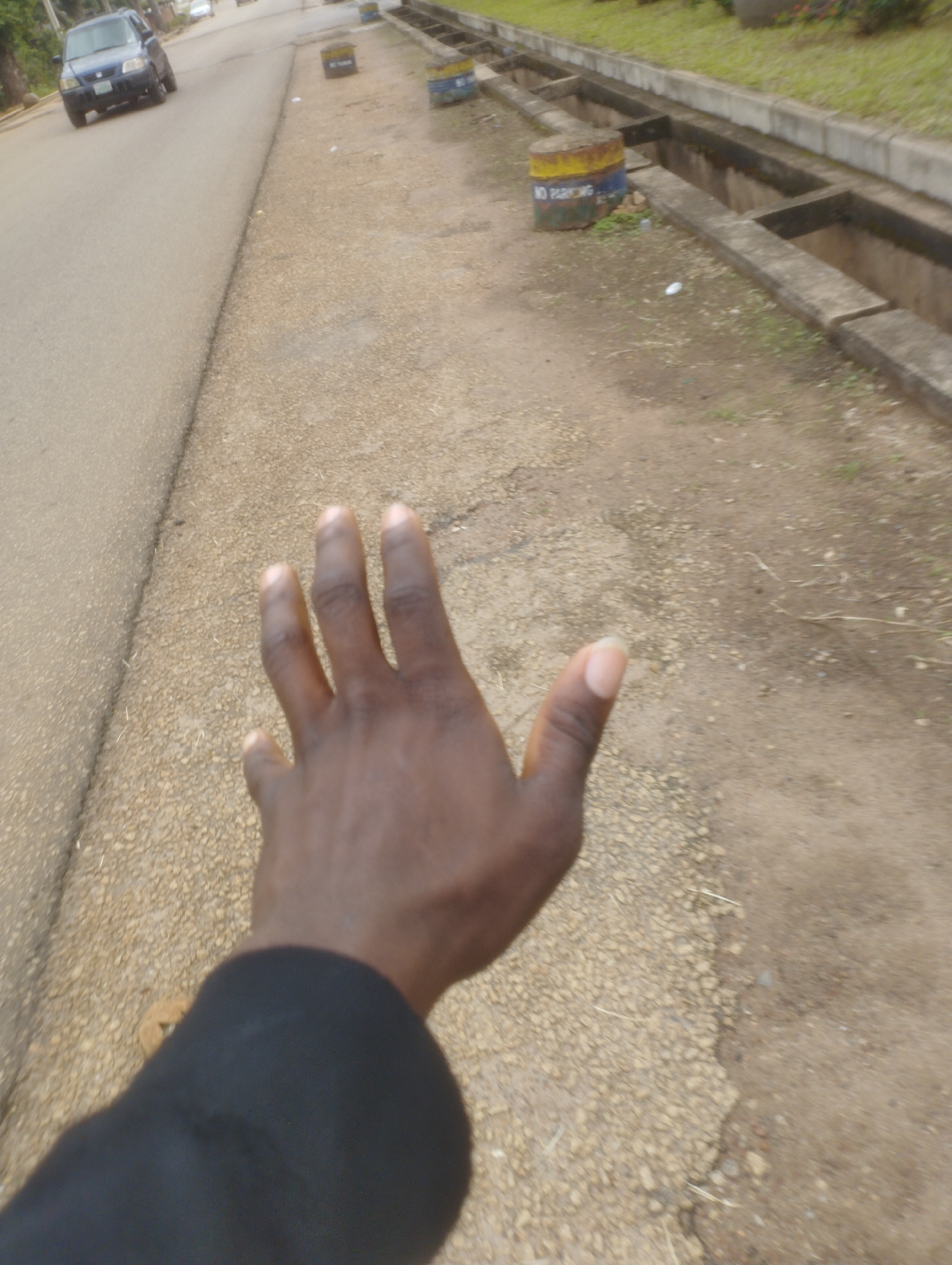 In a mid-level shot, a person's left hand is extended towards the viewer, fingers slightly spread and blurred due to motion. The hand is dark-skinned, with fingernails visible. The person is wearing a black long-sleeved garment. The scene appears to be outdoors, on the side of a road in Jos, Nigeria. The road surface is paved and appears worn. To the right of the road is a gravelly path with scattered dry grass. Further to the right, a concrete drain runs parallel to the road, with a weathered concrete structure on the edge. In the background, a blue sedan is seen driving on the road. Further down the road, several cylindrical objects, possibly street markers or bollards, are visible, one of which is painted with yellow and blue bands and the words "NO PARKING". The overall environment suggests an urban or semi-urban setting, possibly a roadside in Jos.