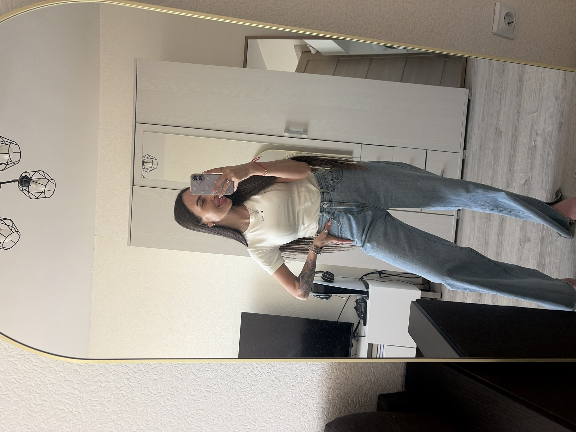 This full-body mirror selfie captures a young woman posing in a modern indoor setting in Saint Petersburg, Russia. The woman, appearing to be in her 20s or early 30s, has long dark hair and a cheerful smile. She is dressed casually in a white short-sleeved t-shirt and light blue, loose-fitting jeans. A red string bracelet is visible on her left wrist, and she wears rings on her fingers. A tattoo is visible on her right forearm. She holds a light-colored smartphone, adorned with a white case featuring light floral patterns, up to capture her reflection.
The background, seen through the mirror, reveals a contemporary room, likely a bedroom or living area. To the woman's left, a white wardrobe or cabinet with light wood accents and silver handles is visible. On her right, a black television or monitor rests on a dark cabinet, next to a white console, possibly a PlayStation 5, with headphones draped over it. The room features light-colored walls, one with a textured pattern and another smooth, complemented by light wooden flooring. A modern ceiling light fixture with black wireframe shades is reflected in the upper left of the mirror, and a white wall socket is visible in the upper right. The reversed text "GENUINE" is partially discernible on her t-shirt. The scene suggests a bright, daytime moment in a well-appointed apartment.