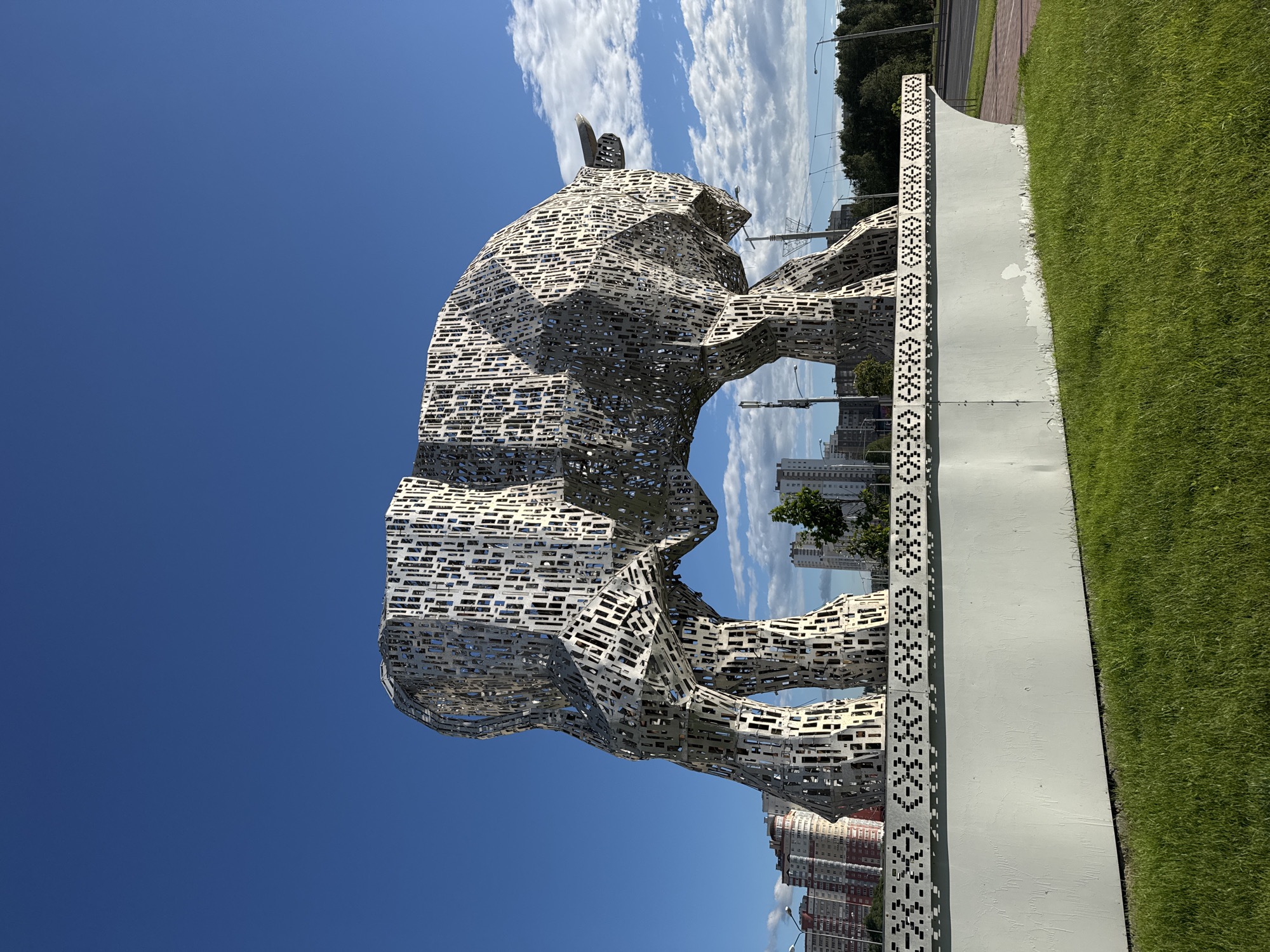 The image shows a large, abstract sculpture of a bull made of metal. The sculpture is located in the city of Minsk, Belarus, specifically at the intersection of Pobediteley Avenue and Privolnaya Street. The bull sculpture is situated on a white, curved pedestal decorated with a black geometric pattern. Behind the sculpture, residential buildings and trees can be seen, with a blue sky and scattered clouds overhead. The ground in the foreground is covered with green grass. The overall scene appears to be an outdoor public space.