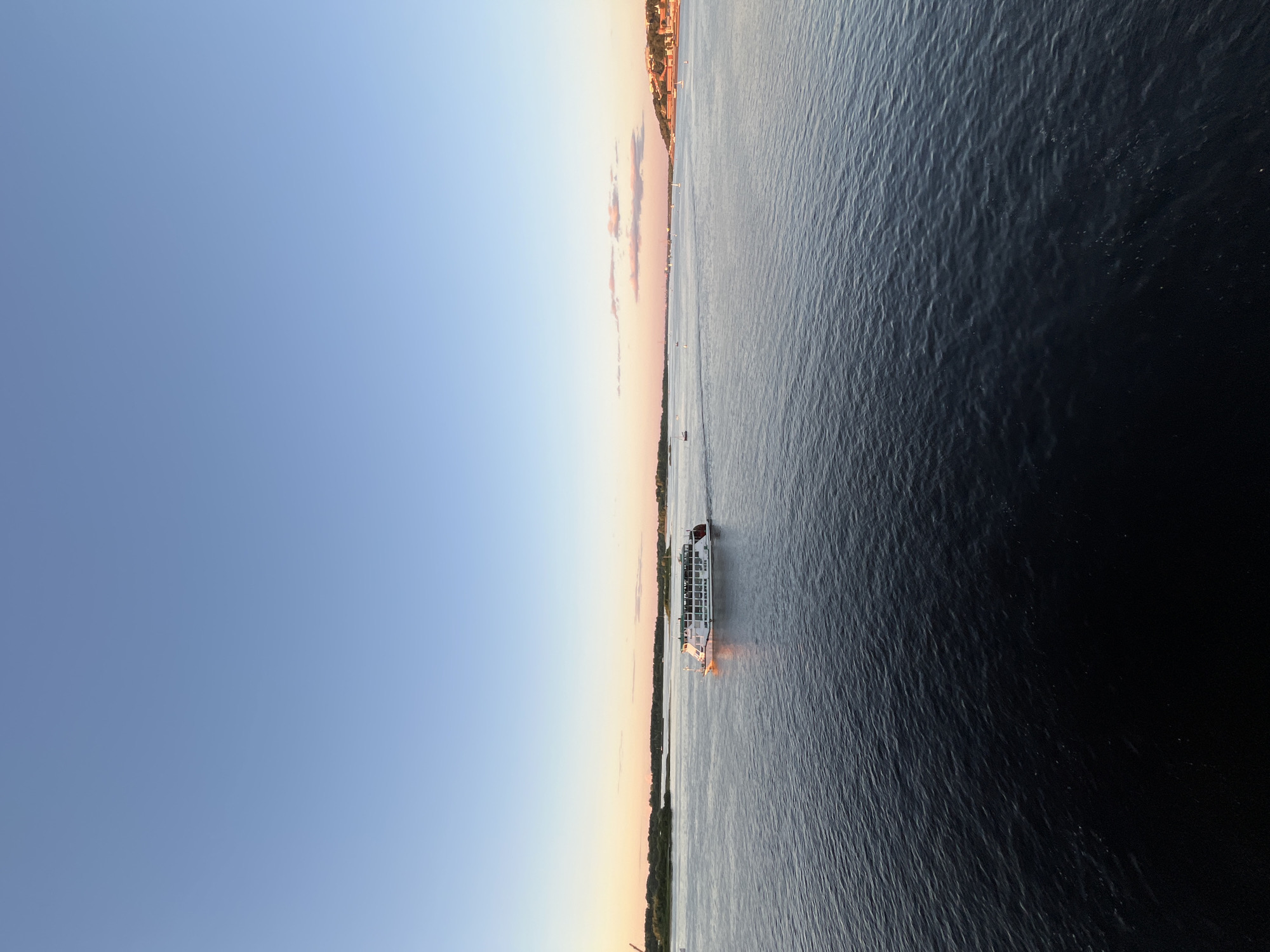 This media file depicts a serene scene of a river at sunset, likely in Nizhny Novgorod, Russia, given the context. The image is oriented vertically, with the sky dominating the left half and the water the right.
The sky is a gradient of soft blues fading into pale oranges and pinks near the horizon, indicating either sunrise or sunset. Scattered, wispy clouds add a touch of color to the sky. Along the horizon, the land is visible, featuring trees and buildings that suggest a populated area. The water is dark and rippling, reflecting the muted colors of the sky.
A white passenger boat is the prominent object in the scene, navigating the river. It has multiple decks and appears to be carrying passengers, although no individuals are clearly discernible. The boat is illuminated by artificial lights, casting a warm glow on the water around it. A few other small boats are visible in the distance on the river, adding to the sense of activity.
The overall mood of the image is peaceful and tranquil, capturing a moment of calm as day transitions into night. The lighting suggests it is late evening. No visible text is present in the image.