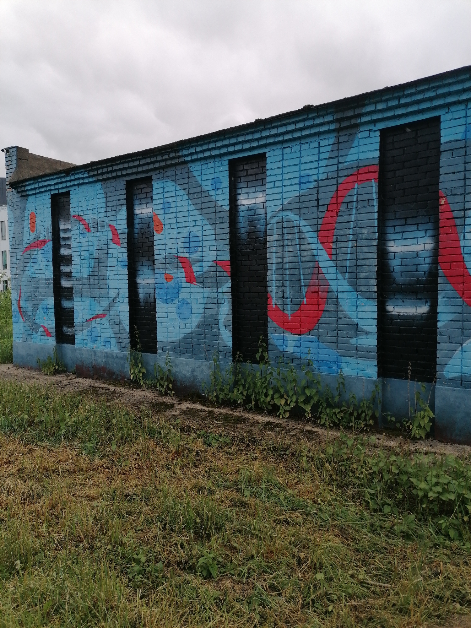 A brick wall with graffiti stands in an urban environment in an unspecified city. The wall is painted blue and black, with red accents in the graffiti. There are four vertical sections that appear to be filled-in window openings, painted black. Weeds and grass grow along the base of the wall, and a patch of dry, yellowed grass extends into the foreground. Overcast sky in the background. A building is barely visible on the left.