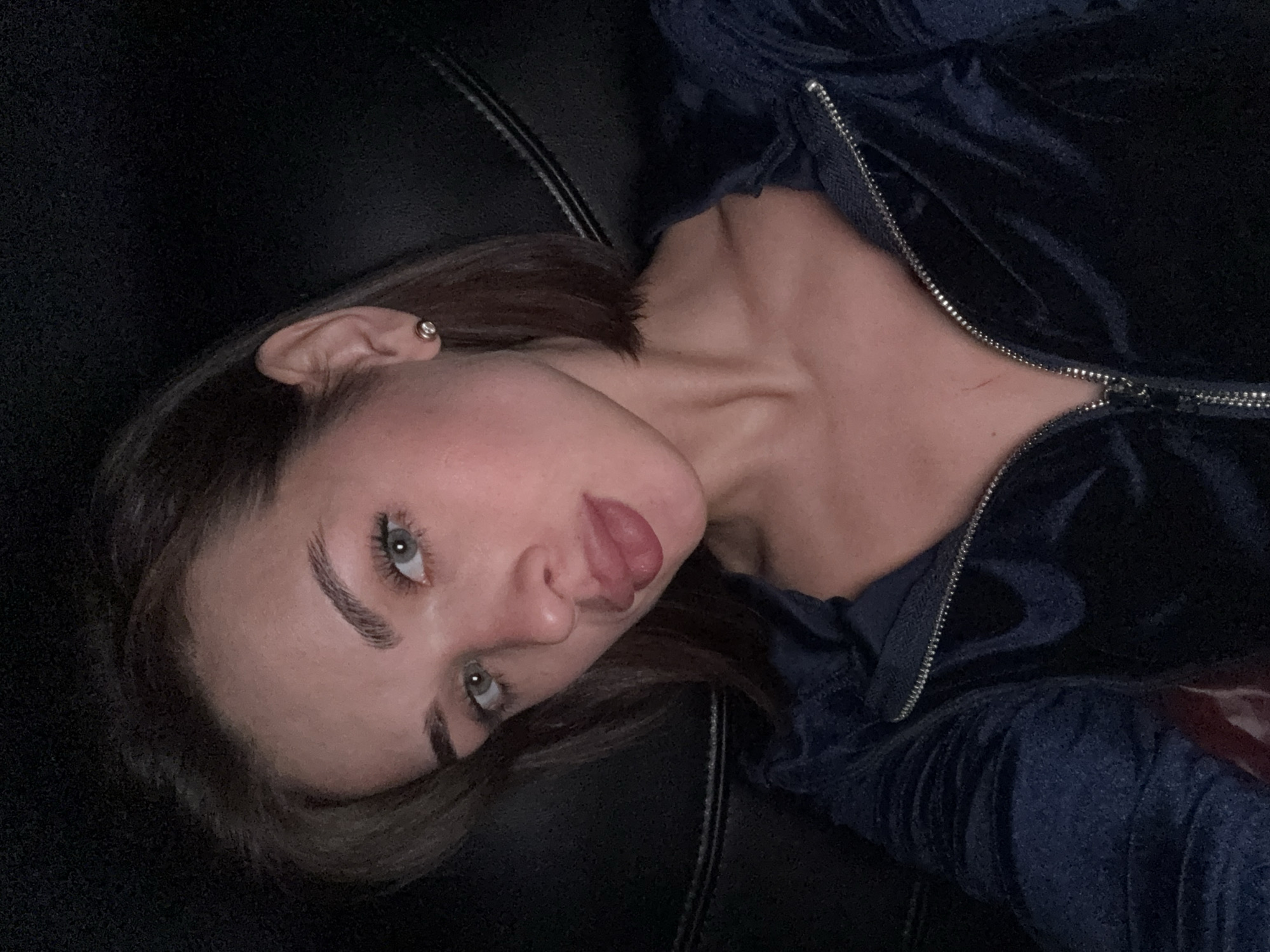 A young woman with light skin and green eyes is lying on a black surface, likely a leather couch or chair. She has brown hair, and her mouth is slightly open, revealing her tongue. She is wearing a dark blue velvet zip-up jacket. The lighting appears to be dim, and the background is dark. There is no information to establish the location is Perm, Russia.