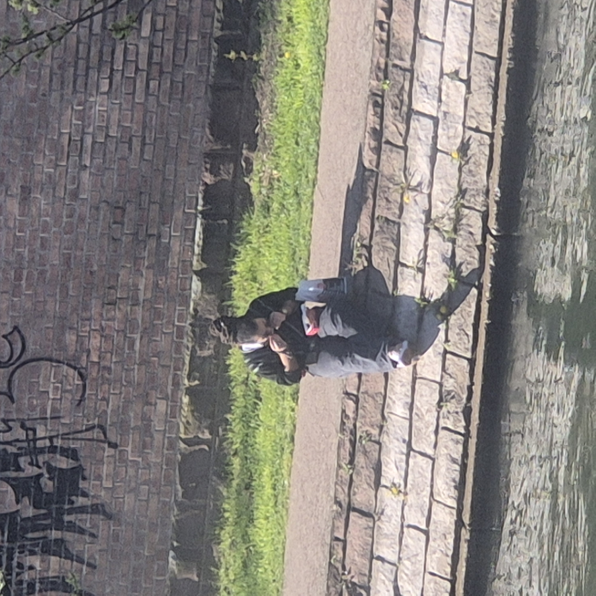 The image shows two people seated on a stone pavement alongside a canal or waterway in Strasbourg, France. They are positioned next to a path that is bordered by green vegetation and a brick wall covered in graffiti. The canal is visible on the right side of the image, with reflections on its surface. One of the individuals is wearing a mask. The overall environment suggests an urban setting adjacent to a natural or semi-natural element.
