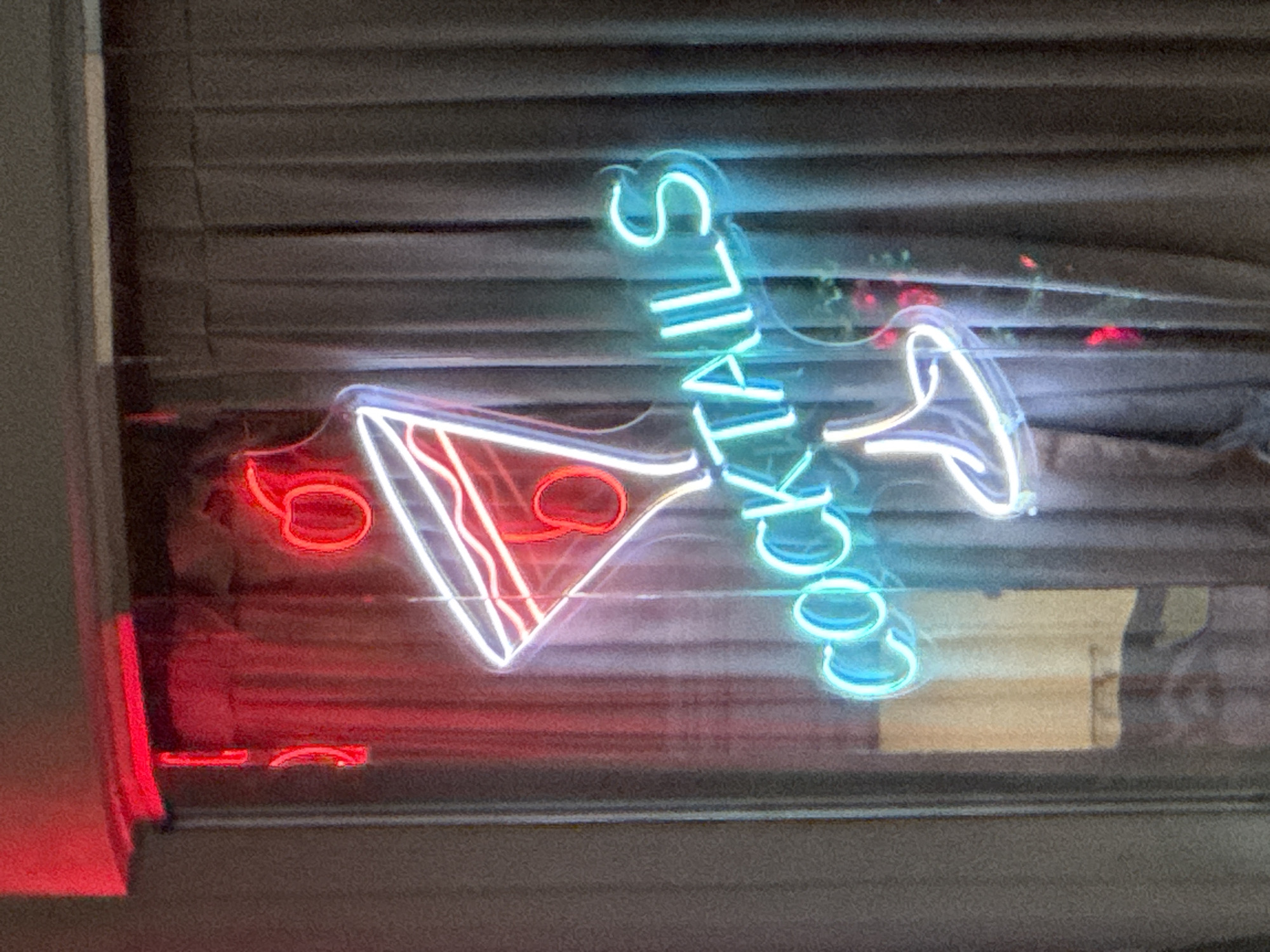 An illuminated neon sign featuring a martini glass with two cherries and the word "COCKTAILS" is displayed in front of a dark gray curtain. The martini glass and the word "COCKTAILS" are illuminated in bright blue and white neon lights. The cherries are depicted in red neon. The sign is positioned in an indoor setting, likely a bar or restaurant, in West Hollywood, United States of America. The lighting is dim, with the neon sign being the primary light source. The curtain appears to be made of a heavy fabric and is partially obscuring a window or wall behind it.