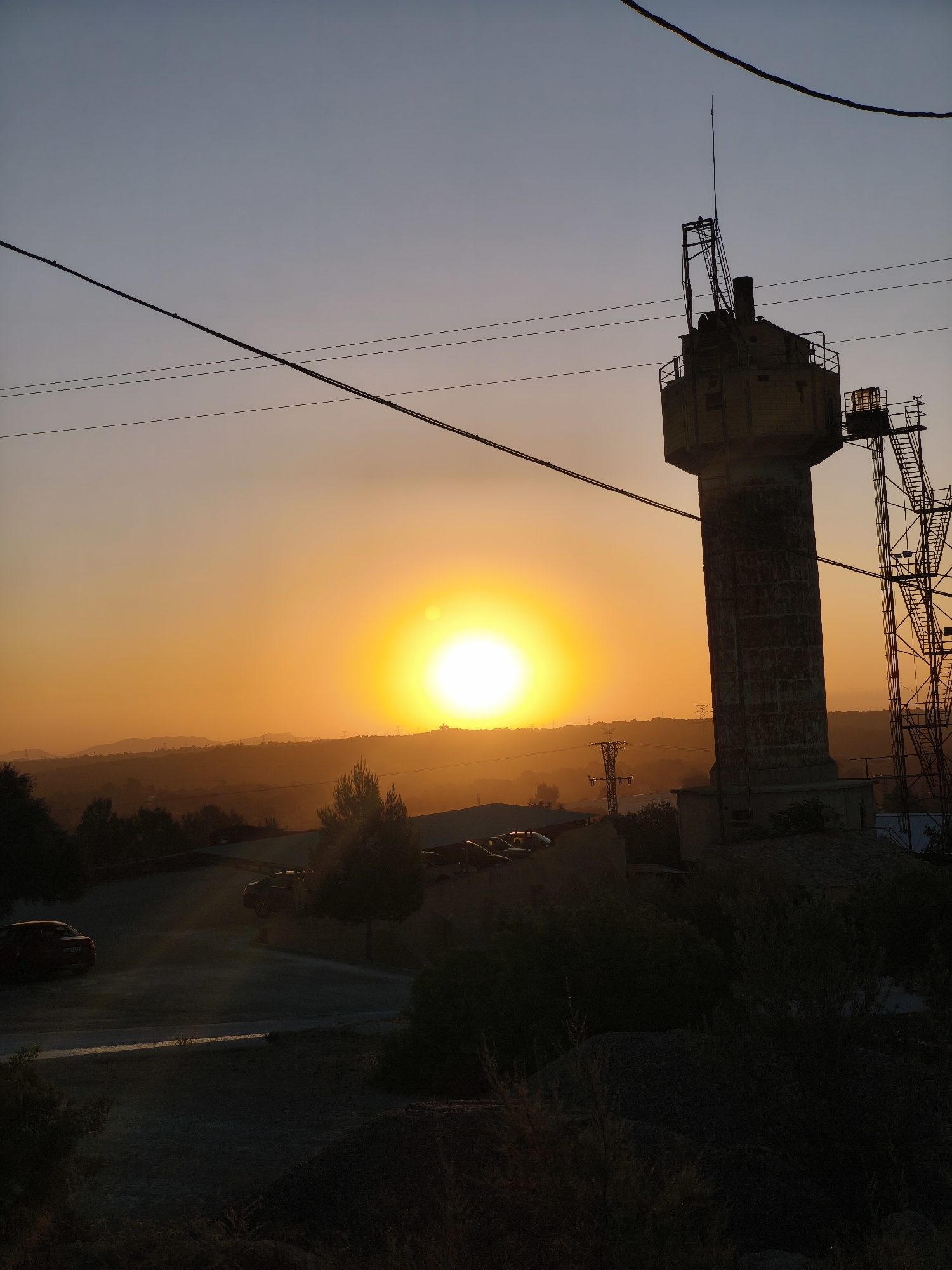 This image captures a sunset in Riba-roja de Túria, Spain, with a prominent water tower silhouetted against the bright sun. The sky transitions from a deep blue at the top to a warm orange near the horizon. In the foreground, a road curves through a sparsely vegetated area, with a red car parked on the left. Several trees dot the landscape, some appearing silhouetted. A series of power lines stretch across the sky. To the right, the tall, concrete water tower stands with an antenna on top, accompanied by a complex metal structure. Further in the background, rolling hills and distant structures are visible, softened by the sunset's glow. The overall scene conveys a sense of twilight stillness in an industrial or semi-rural setting.