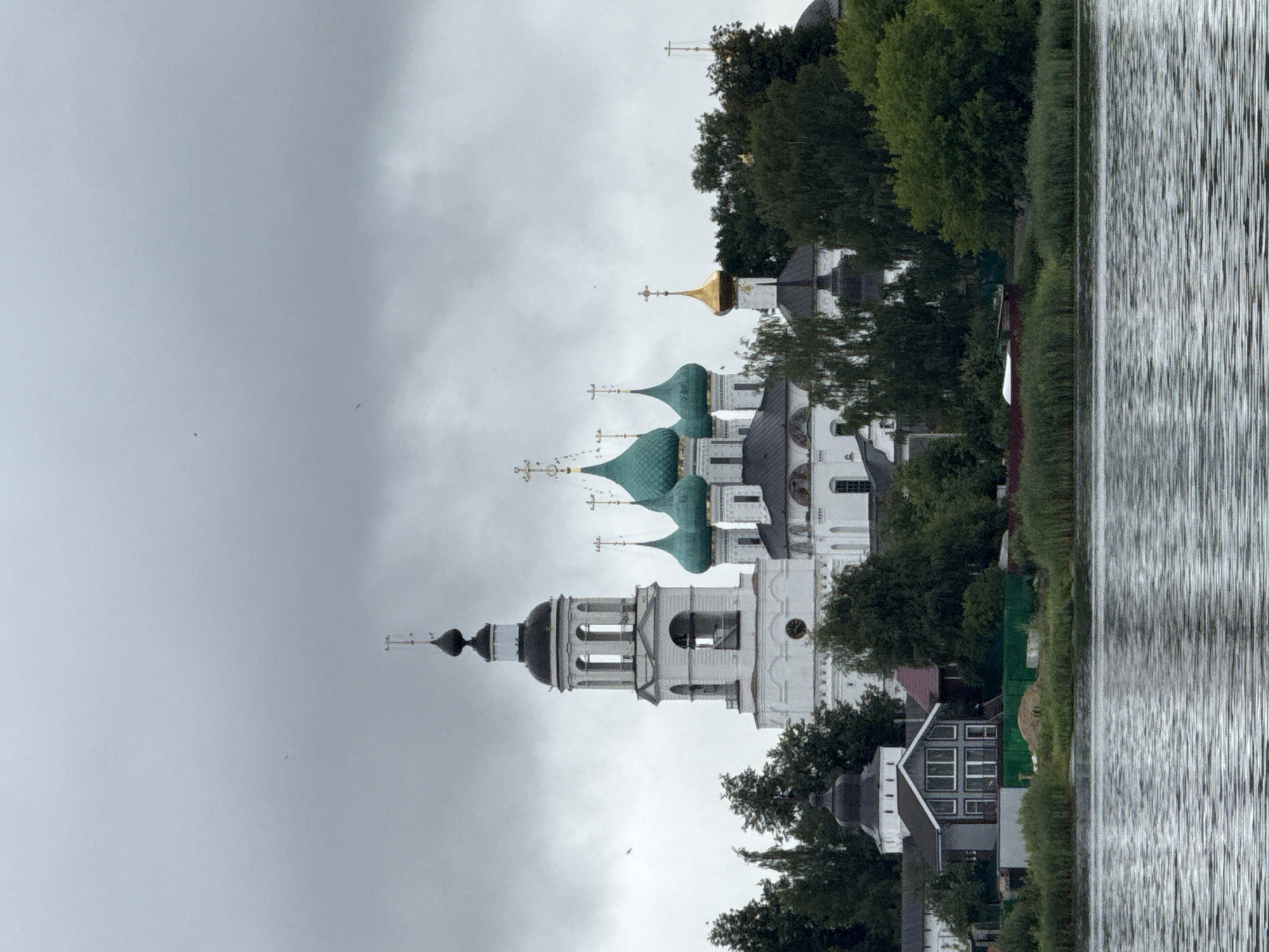 This is a distant, medium shot of a monastery complex on the bank of a body of water, under a cloudy sky. The monastery features several white buildings with distinctive green and gold onion domes, topped with crosses. A tall white bell tower with a black dome stands prominently to the left of the main complex. In the foreground, the rippling surface of the water is visible, reflecting the overcast sky. Behind the monastery, dense green trees and vegetation line the shore. To the left, a small cluster of residential buildings with dark roofs is situated between the monastery and the water. The overall scene suggests a serene, perhaps historic, location.