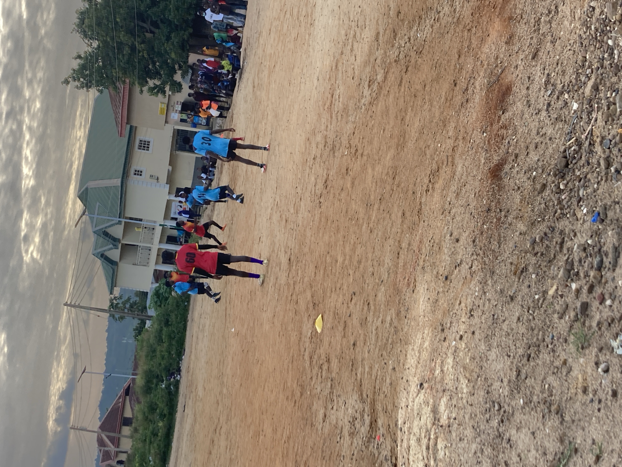 The image depicts an outdoor scene with a group of young men playing soccer on a dirt field, likely in Jos, Nigeria, based on the architecture and attire. The sky is overcast, suggesting a cloudy or late afternoon setting. In the background, several buildings are visible, characterized by light-colored facades, multiple stories, and green or red-tiled roofs, typical of urban or semi-urban structures in the region. Some of these buildings appear to be houses or small commercial establishments. A number of spectators are gathered on the sidelines, watching the game. The field itself is unkempt, with patches of dry grass and scattered debris, including a yellow marker cone in the foreground. The players are wearing various colored jerseys, with one prominent player in a red jersey with the number "60" on the back, and another in a blue jersey with the number "30". The overall impression is one of informal sporting activity within a residential or mixed-use urban environment.