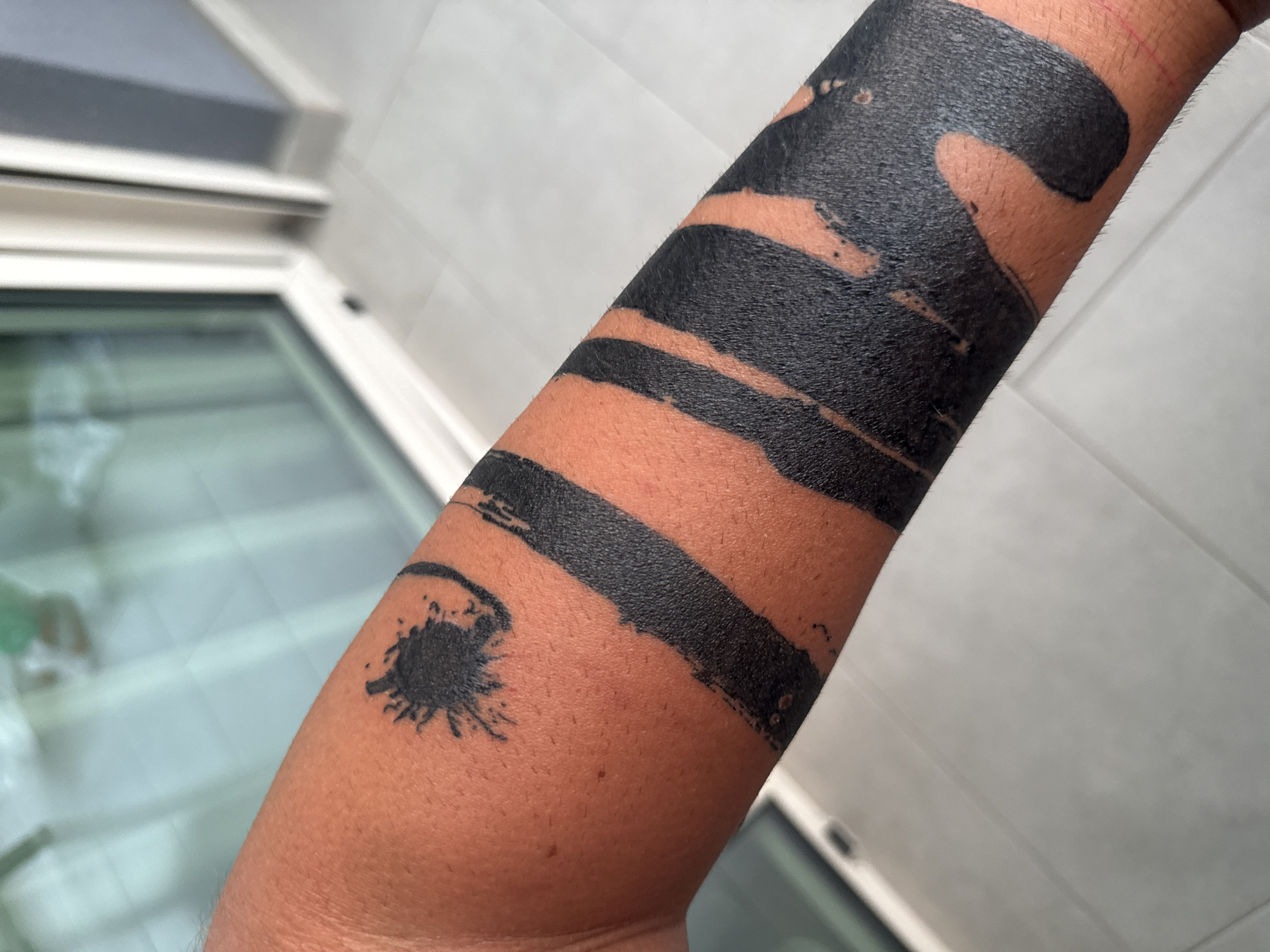 This media file shows a close-up view of a person's arm with a black tattoo. The tattoo is abstract, featuring broad brushstrokes and splatters of black ink. Some areas of skin are visible between the tattooed sections, showing a light to medium skin tone. The arm appears to be in the process of healing or is freshly tattooed, as there are some minor blemishes and what might be fine hairs visible on the skin. The background is blurred, but suggests an indoor setting with white tiled walls and a window. The lighting appears to be natural and bright, indicating it is daytime. No text is visible in the image. The overall impression is one of a detailed focus on the tattoo itself, highlighting its texture and design.