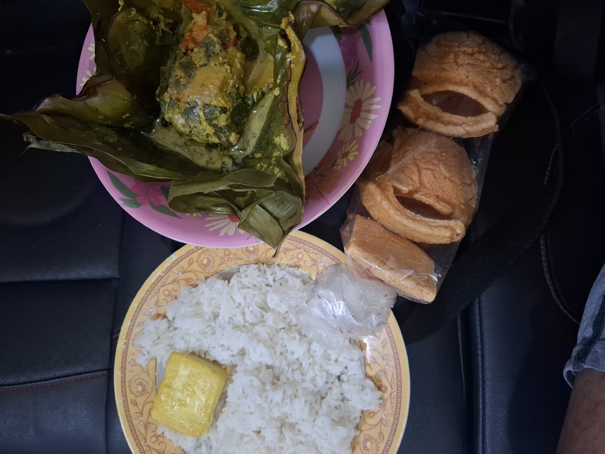 The image shows a meal served on plates, likely taken in a car given the dark leather upholstery visible in the background, suggesting a portable dining setting. The meal consists of white rice, a piece of fried tofu, and a dish of fish or chicken wrapped in banana leaves, served in a yellow-patterned plate and a pink-patterned plate respectively. There are also several pieces of fried crackers in a plastic bag. The scene is dimly lit, indicating it might be inside a vehicle with the engine running or parked, possibly in Maleber, Indonesia, where such a portable meal might be consumed. There are no people clearly visible in the frame.