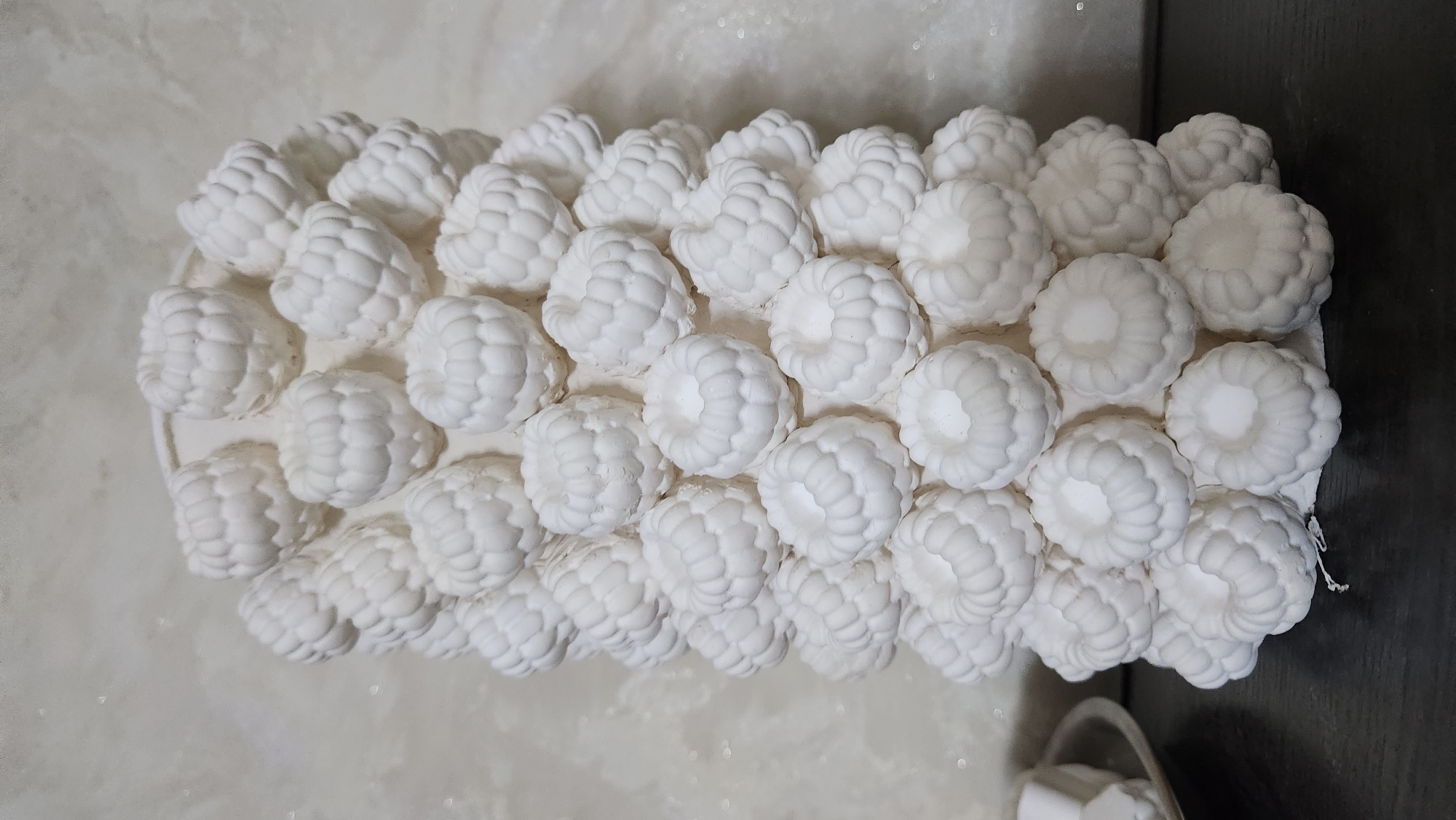The image shows a white, cylindrical object covered in what appears to be a cluster of small, round, textured elements resembling raspberries. The object is standing upright against a textured, light-colored wall. The bottom of the object rests on a dark surface. To the left of the object, a small portion of a silver dish holding some white, rectangular items is visible. The lighting is soft, with some glare on the wall. There are no people or animals present in the image. The location of the city of Buynaksk, Russia, is not depicted or relevant to the visual content of the image.
