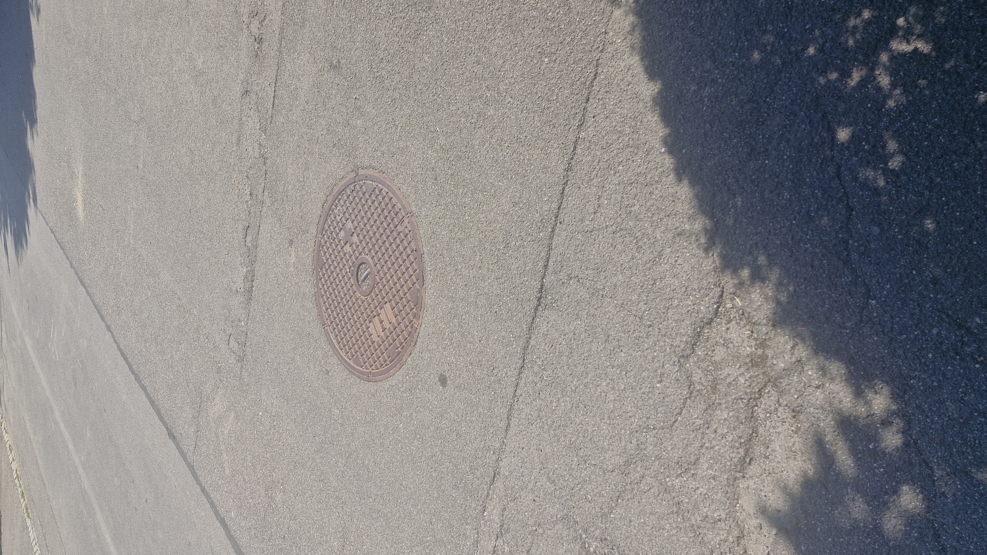 The image is a close-up, eye-level shot of a manhole cover on a paved street in Wattenwil, Switzerland. The manhole cover is round and made of metal, with a grid-like pattern and some text or symbols visible. The asphalt of the road is gray and shows some signs of wear, including cracks. In the lower part of the image, a dark shadow is cast, likely from trees or a building, with dappled light filtering through. The upper portion of the image shows more of the road, with a faint line indicating a lane divider. The lighting suggests it is daytime, with sunlight creating the shadow.