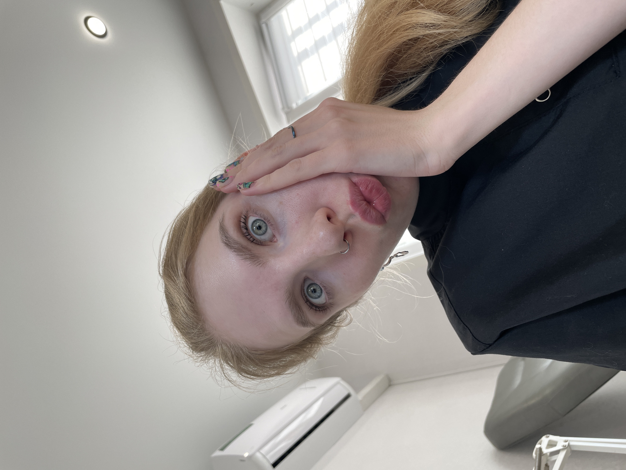 A young, fair-skinned person with blonde hair and blue eyes is positioned in the center of the image, seemingly taking a selfie. They are wearing a black top and have a ring on their finger. Their hand partially covers their face, and their lips are pursed. A nose ring is visible. The background includes a white wall, a window with white curtains, an air conditioning unit, and a grey object in the lower right corner that could be a piece of furniture. There is a light fixture on the ceiling. There is no information available to confirm the location as Izhevsk, Russia.