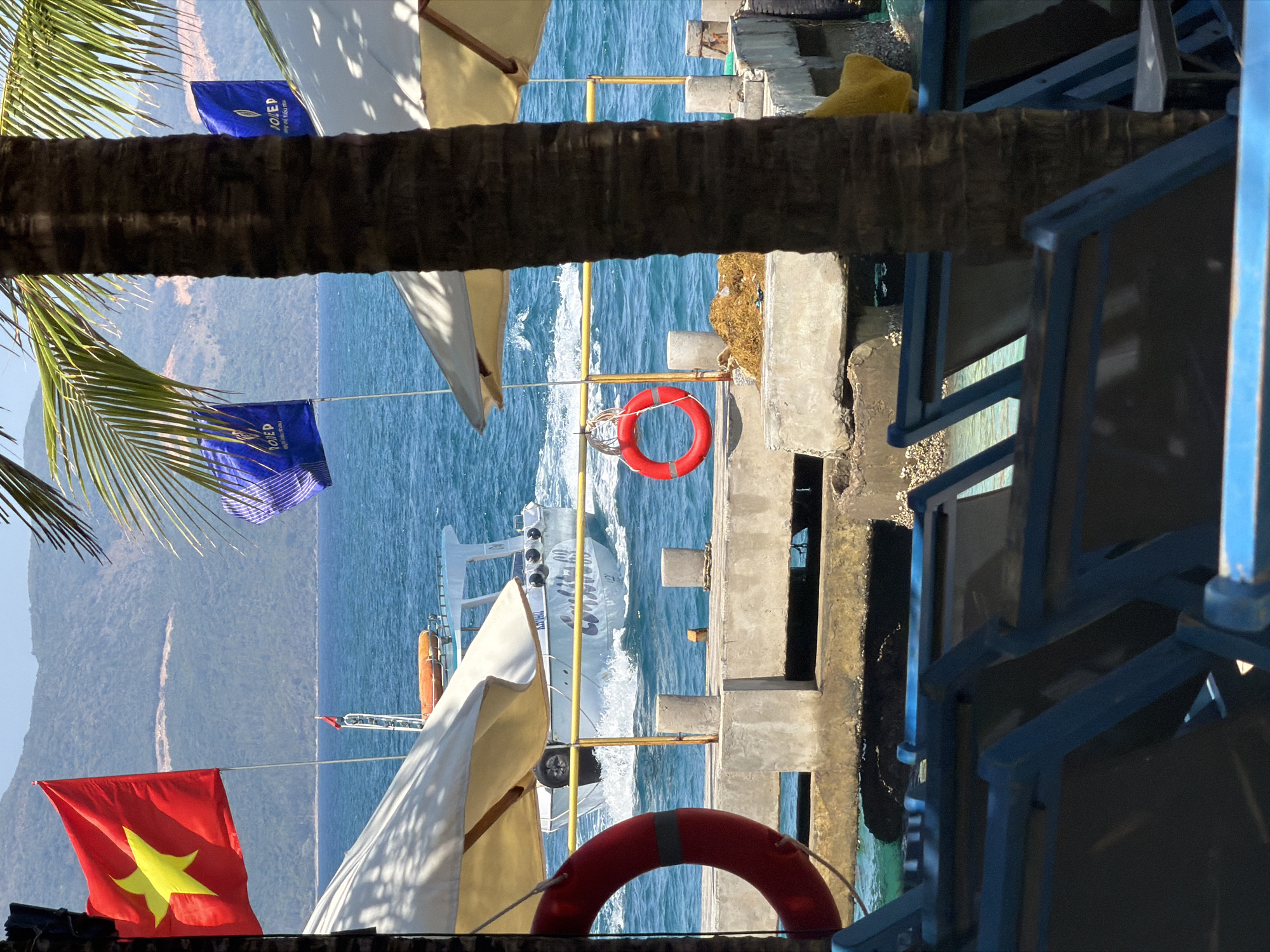 The image shows a waterfront scene in Nam Nha Trang Ward, Vietnam. A boat is traveling on the blue water. The shore is lined with concrete structures, and there are blue chairs on the right. A palm tree trunk is visible in the foreground. There are red and blue flags visible. A Vietnamese flag is also visible on the lower left of the image. Red life preservers hang along the dock.