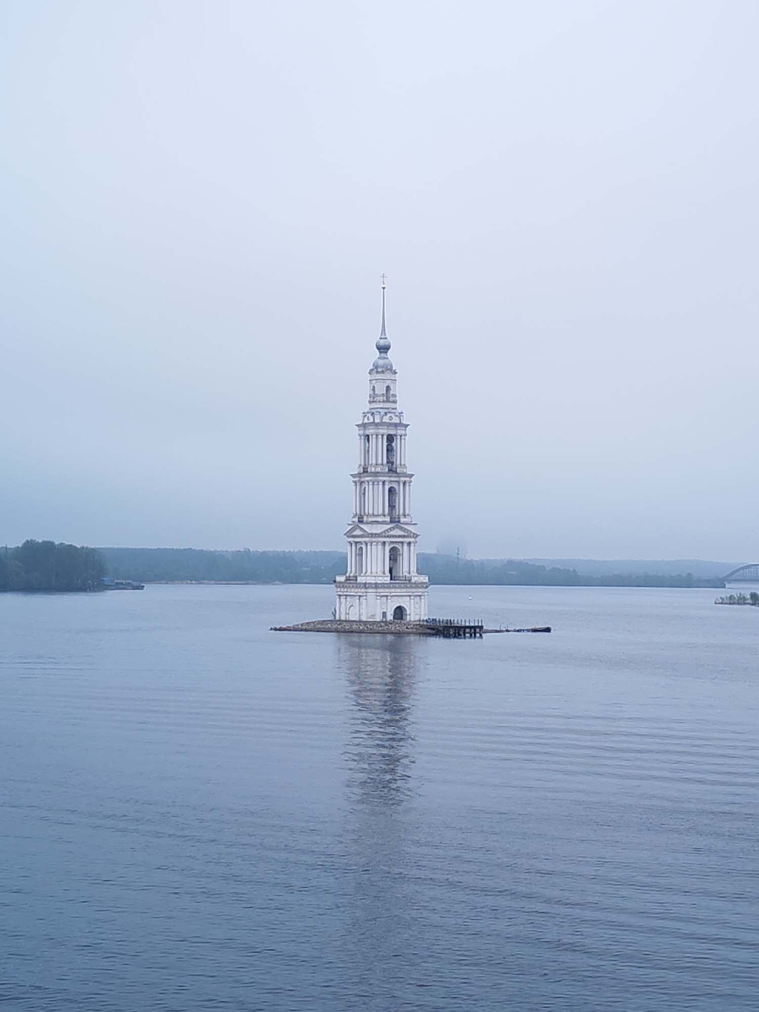 The image depicts the flooded bell tower, known as the Kalyazin Bell Tower, standing alone in the waters of the Uglich Reservoir on the Volga River in Kalyazin, Russia. The bell tower is tall, white, and tiered, with arched openings on each level. A small, dark-colored dock or platform extends from the base of the tower into the water. The water is calm and reflects the tower, creating a mirrored image. The sky is overcast and grey, casting a muted light on the scene. In the background, across the water, there is a hazy shoreline with trees and some structures, including a bridge visible on the right side of the frame. A barge or similar vessel is seen moored closer to the shoreline on the left. There are no people visible in the image.