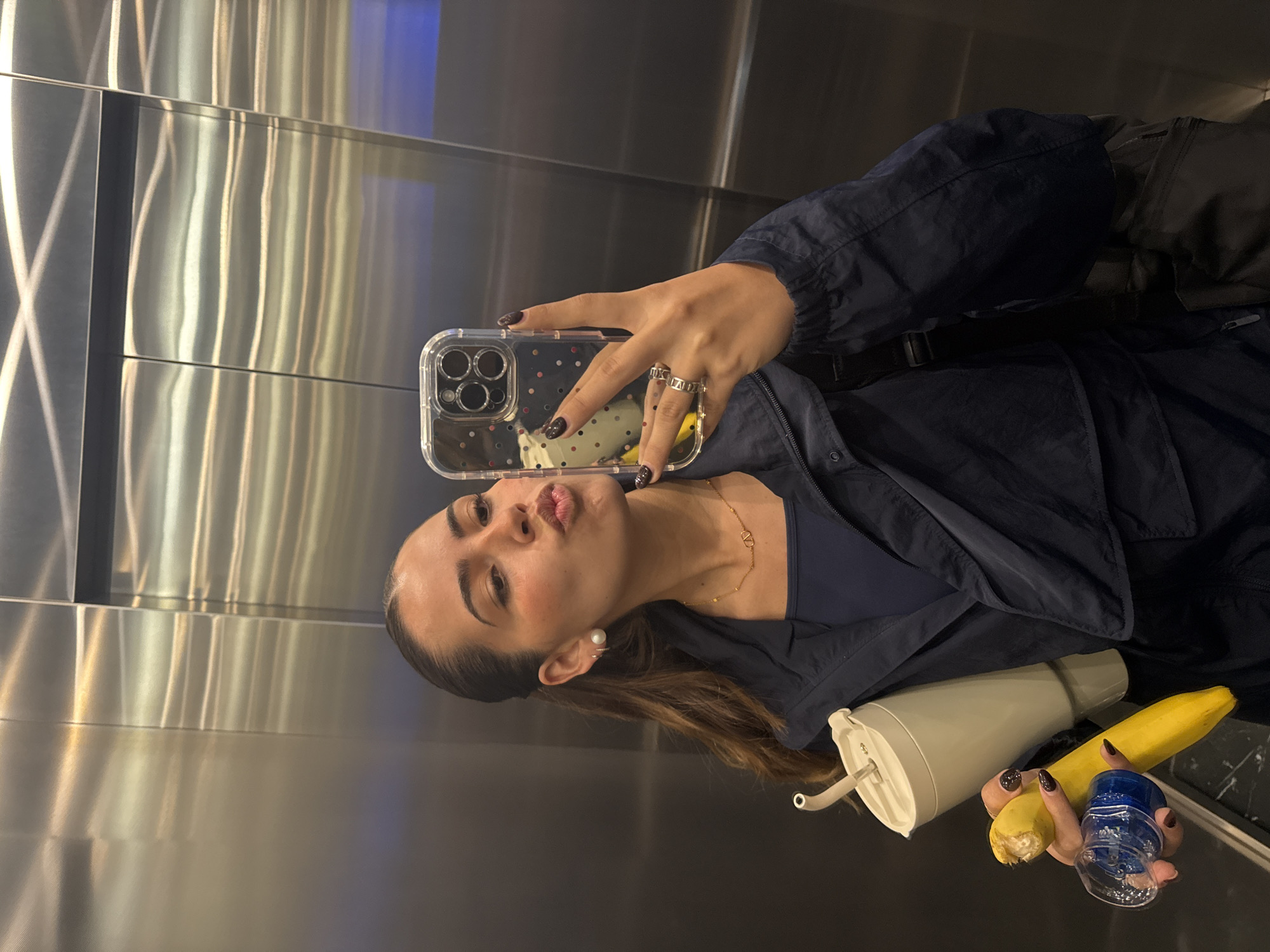 A young woman takes a selfie in a reflective, metallic-walled interior, likely an elevator or a modern building, in Puebla, Mexico. She is seen from the chest up, with her head tilted and a playful, pouting expression. Her hair is pulled back, revealing a pearl stud earring and an ear cuff on her left ear. She wears a dark blue long-sleeved top or jacket over a darker inner layer, accessorized with a delicate gold chain necklace featuring a 'V' shaped pendant, possibly a Valentino logo. Her fingers are adorned with multiple rings, one of which appears to have Roman numeral engravings, and her nails are painted a dark shade.
She holds a smartphone with a clear, polka-dotted case to capture the reflection. Near her arm, she is also holding a bright yellow banana and a small, translucent blue container or cup. A light grey or beige reusable water bottle with a straw is visible beside these items. The background consists of vertically paneled, highly polished metallic surfaces that reflect light in streaks and patterns, indicating an indoor, contemporary environment with artificial lighting.