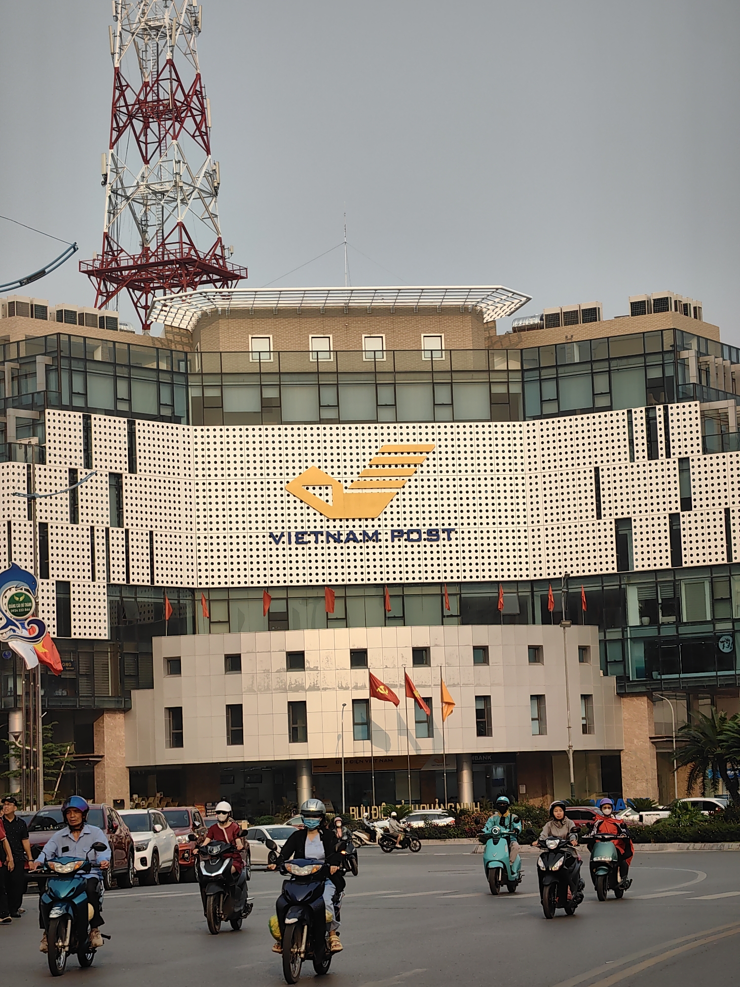 The image depicts a street scene in Hong Gai Ward, Vietnam, in front of a Vietnam Post office building. The building is modern, with glass and white panels, and features the Vietnam Post logo prominently. A red and white telecommunications tower rises behind the building. The street is busy with motorbikes, typical of Vietnam. Several riders are visible, wearing helmets and casual clothing. Some cars are also parked along the street. Flags, including Vietnamese flags, are displayed in front of the building. The lighting suggests it is late afternoon or early morning.