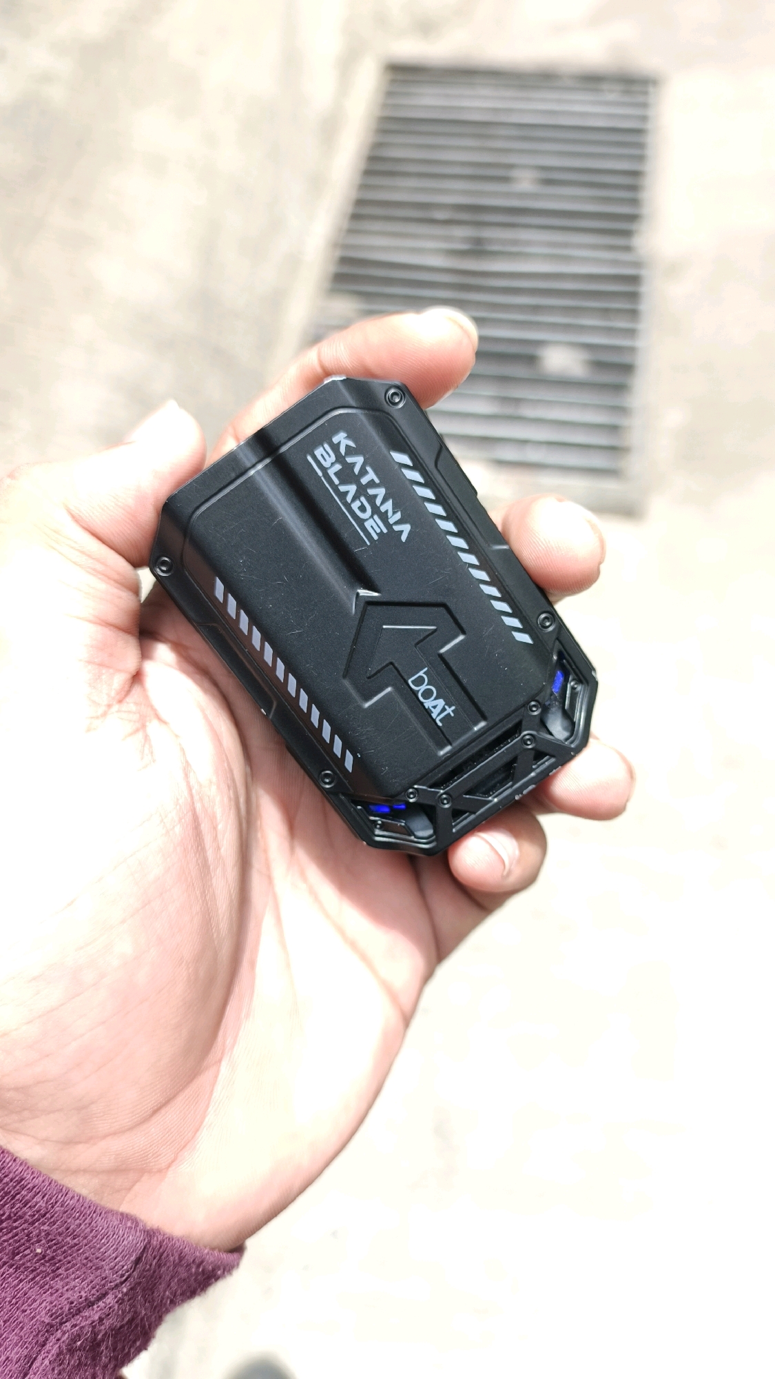 The image shows a person's hand holding a black portable charger with the brand name "boat" and "KATANA BLADE" printed on it. The charger has a rugged design with silver accents and some blue LED lights visible on the side. The background appears to be an outdoor setting with a metal grate and a concrete surface, suggesting a street or sidewalk in Mumbai, India, possibly near a drain. The lighting indicates it is daytime.