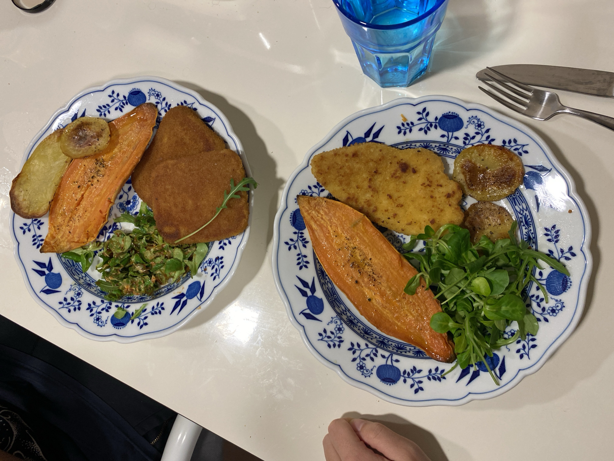 Two plates of food are arranged on a white surface. Each plate has a blue and white floral pattern around the rim. The plate on the left contains two rectangular pieces of golden-brown food, a slice of cooked sweet potato, a potato slice, and some greenery. The plate on the right contains a piece of golden-brown food, a slice of cooked sweet potato, several potato slices, and greenery. A blue glass, a fork, and a knife are located near the right plate. A person's hand is partially visible near the bottom right of the image. The environment appears to be indoors, likely a dining area. The location is not identifiable as Villaviciosa, Spain from this image.