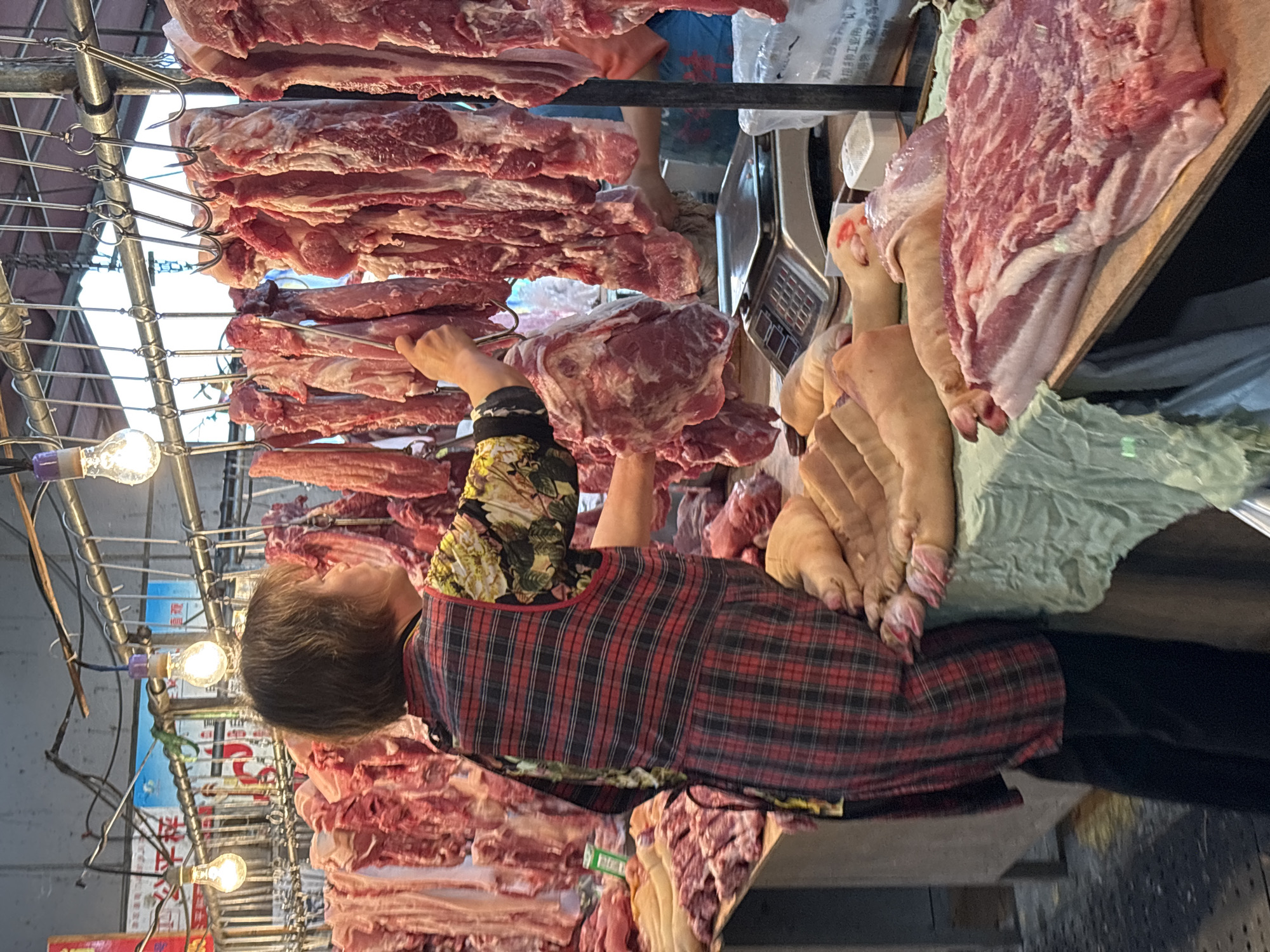 In Jiangjin, China, a middle-aged woman with short gray hair stands in a butcher shop, wearing a red and black plaid apron over a floral-sleeved shirt, with black pants. She reaches up to handle a large cut of raw meat hanging from a metal rack. The shop is filled with various cuts of pork, some hanging and some displayed on a counter, including several pig feet. A digital scale sits on the counter alongside the meat. The shop is brightly lit with multiple light bulbs.