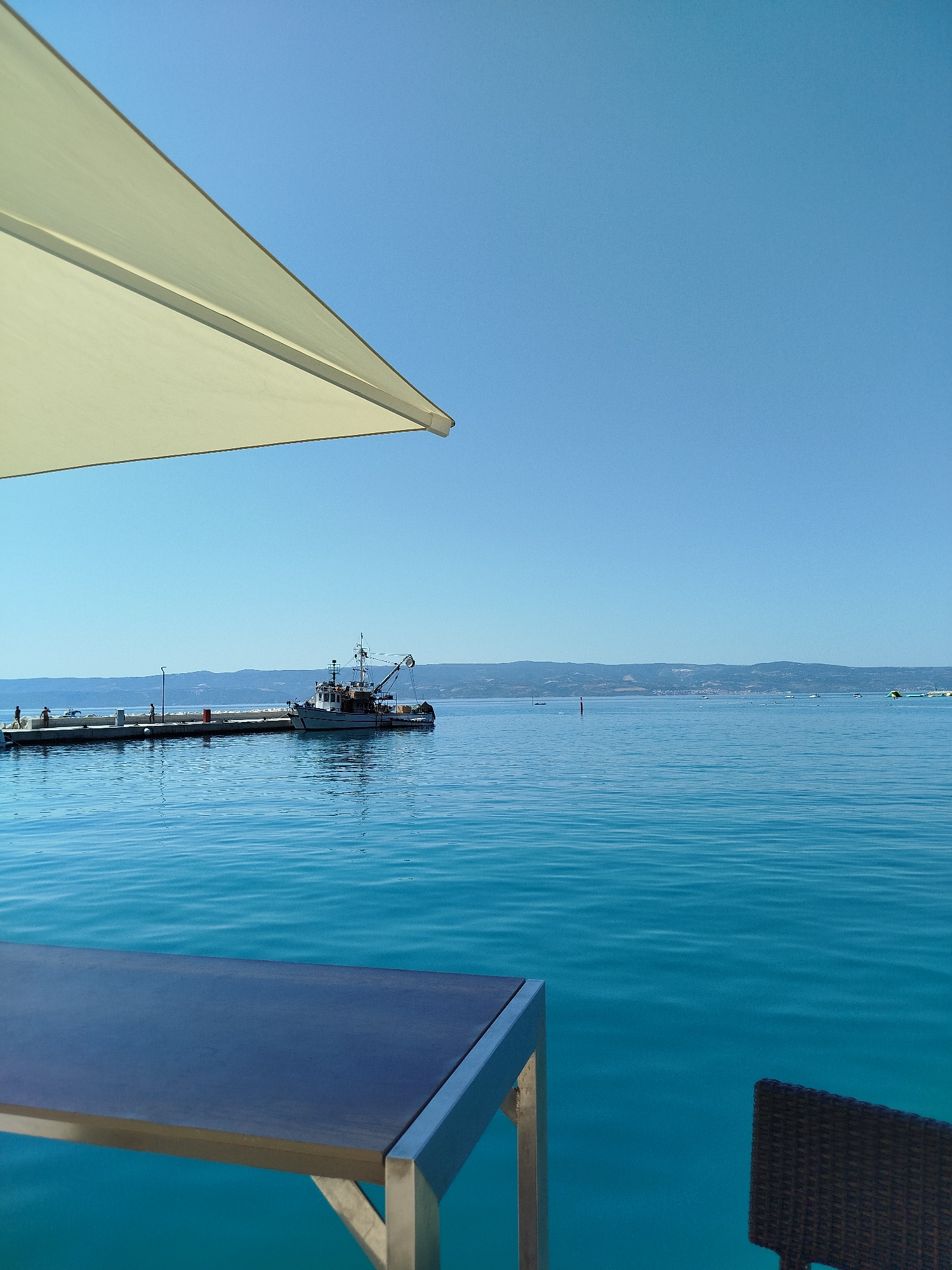 A wide-angle, eye-level, outdoor, daytime shot captures a tranquil scene by the clear turquoise Adriatic Sea in Omiš, Croatia. In the foreground, a dark-colored table with a metallic frame sits partially in view, suggesting a cafe or restaurant setting. To the right, a dark wicker chair back is visible. A large, light-colored umbrella extends from the upper left corner, casting a shadow that partially obscures the scene.
In the middle ground, a long concrete pier juts out into the sea. A fishing boat with a white hull and superstructure is moored alongside the pier. Several small figures, appearing to be people, are discernible on the pier, with at least two standing near the boat and others further down. The water exhibits gentle ripples, reflecting the clear, cloudless, bright blue sky.
The background features a distant, hazy mountain range stretching across the horizon. A few scattered boats and buoys are visible on the water in the far distance. The overall impression is one of a peaceful, sun-drenched coastal location.