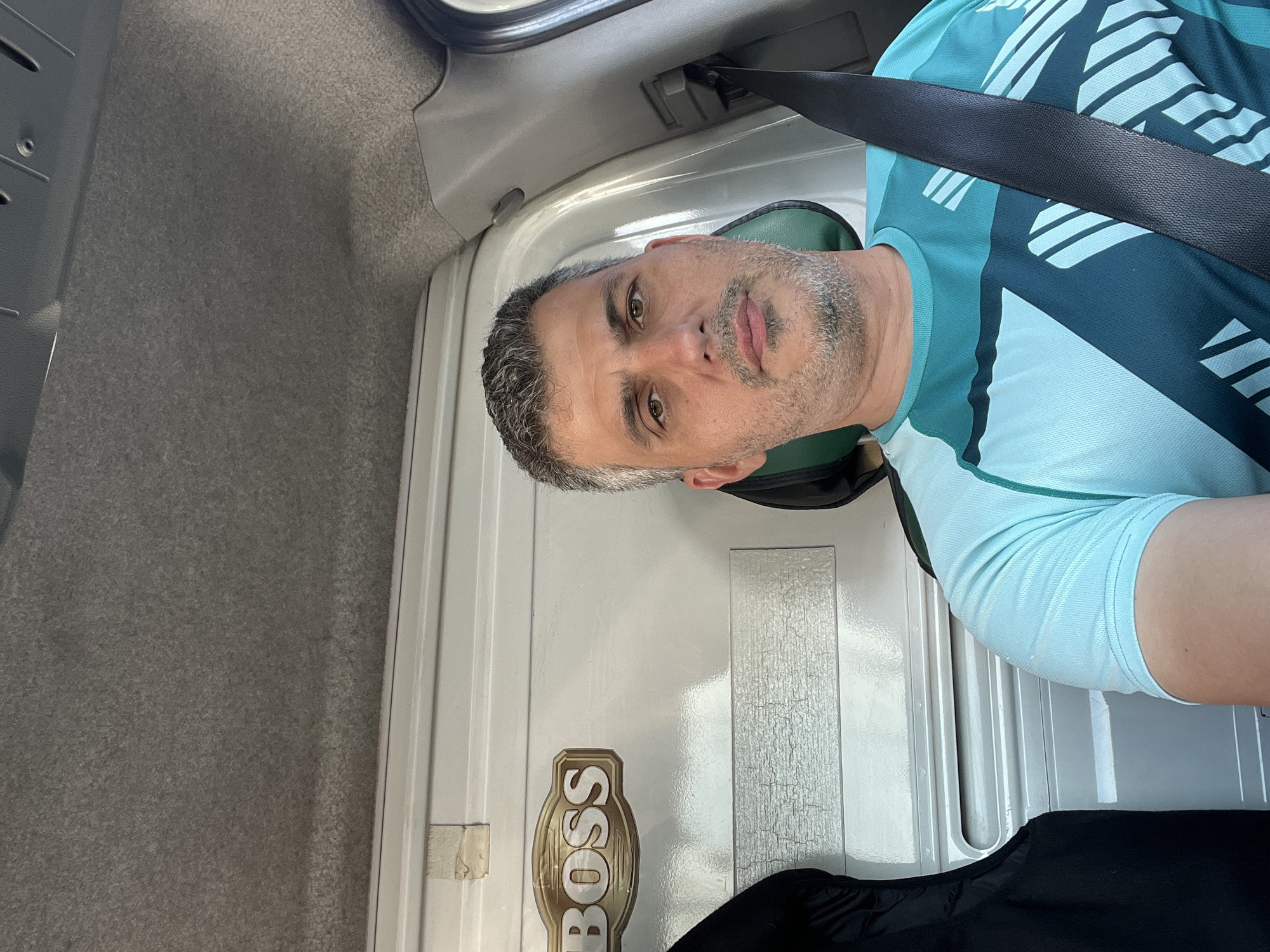 A middle-aged man with grey hair and a beard is sitting in the driver's seat of a vehicle, looking directly at the camera. He is wearing a teal and dark green athletic shirt and a black seatbelt is fastened across his chest. The background appears to be the interior of a van or truck, with a white wall and a "BOSS" logo visible on the left. The ceiling of the vehicle is lined with grey fabric. The scene is captured from a slightly low angle, looking up towards the man's face. There is no indication of the specific city or location.