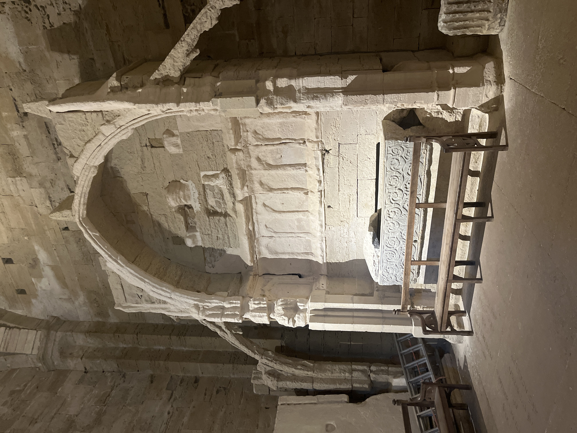 The image depicts the interior of a stone structure, likely an ancient church or cathedral, in Villeneuve-lès-Maguelone, France. The central focus is a large, ornate stone sarcophagus situated within a recessed alcove. The sarcophagus is carved with intricate patterns and rests on a stone base. In front of the sarcophagus is a long, wooden bench. To the left of the sarcophagus, a ladder is leaning against a stone wall, and another wooden chair is visible. The walls and ceiling are constructed from rough-hewn stone blocks, characteristic of historical architecture. Arched structures and carved stone details are evident throughout the space. The lighting is dim, suggesting an indoor environment with artificial illumination. The floor is made of stone or concrete.