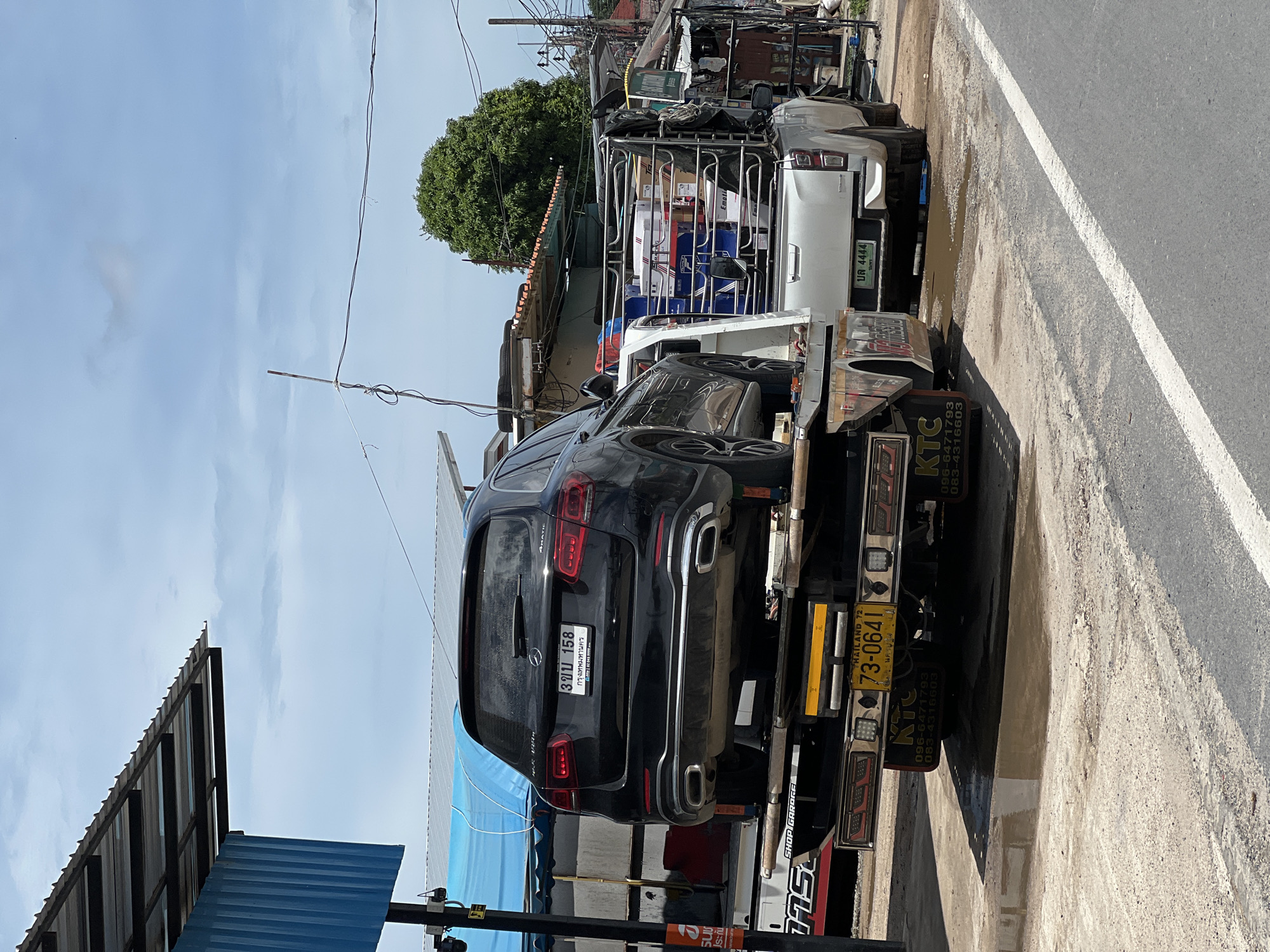 A black Mercedes-Benz SUV is being transported on the back of a white tow truck on a paved road in Baan Bang Rak, Thailand. The SUV, with license plate 3 จบ 158, is positioned on the tow truck's flatbed. Behind the tow truck, a silver pickup truck is loaded with boxes. The scene is outdoors under a cloudy sky, with buildings and a tree visible in the background. There are puddles of water on the ground near the vehicles. The tow truck has yellow and red lights on its rear. The license plate on the tow truck reads 73-0641. The pickup truck has a license plate that reads บจ 4444.