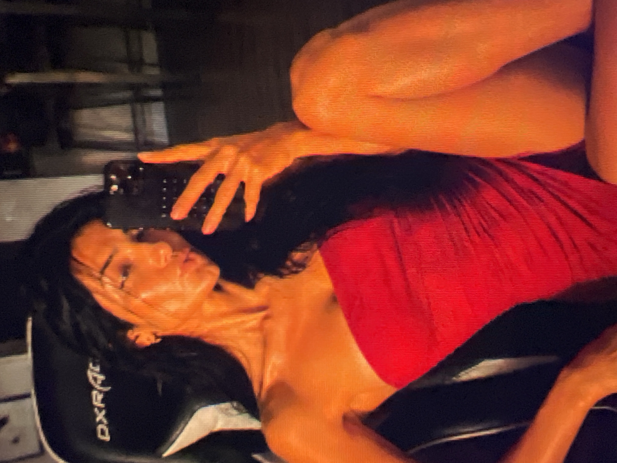 A woman with long, dark hair is sitting in a black gaming chair and taking a selfie with her phone. She is wearing a strapless red dress. The chair has "DXRacer" written on it in white lettering. The background is dark, with some blurry furniture visible. The lighting in the image is warm and appears to be artificial.