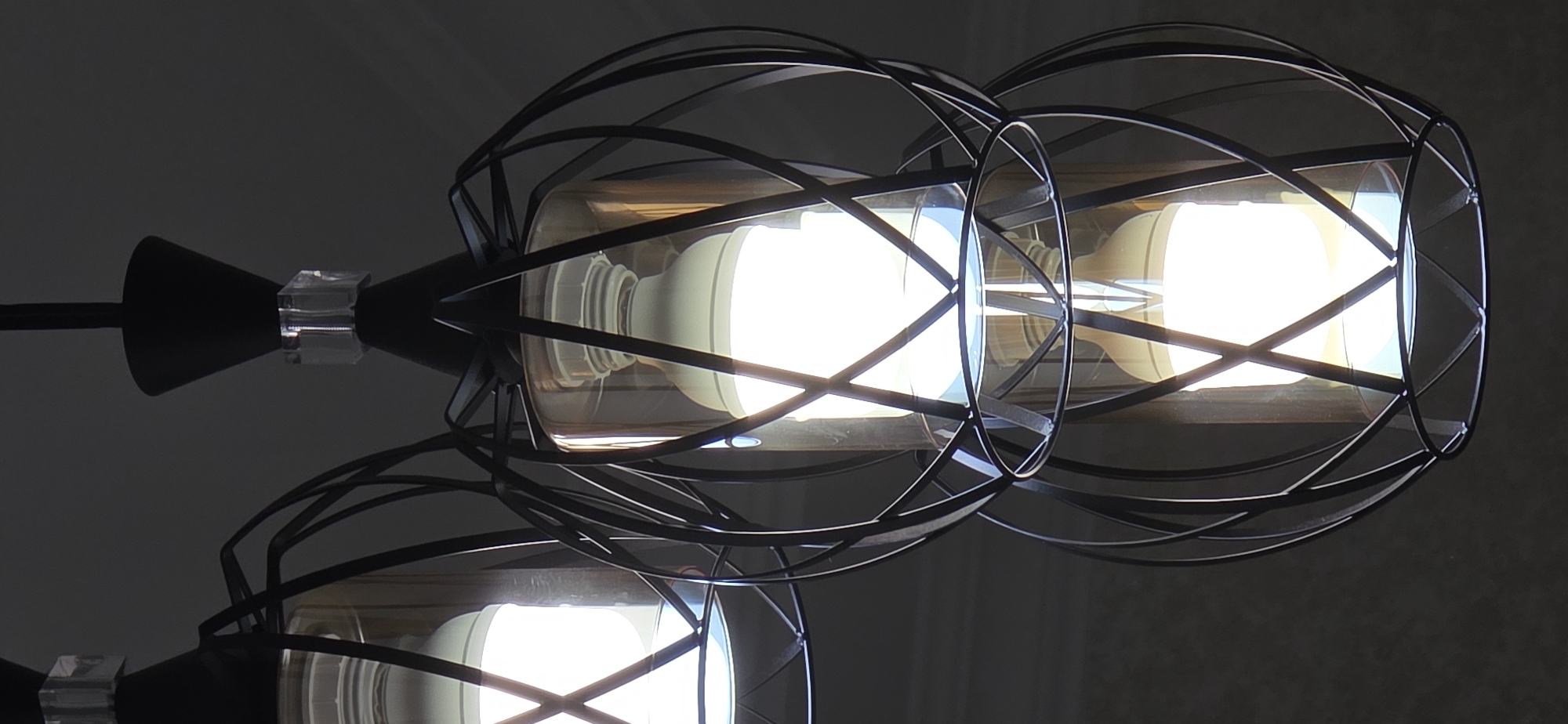 The image shows a close-up of a modern chandelier with three light fixtures. Each fixture consists of a black metal cage with an intricate, geometric design, enclosing a clear glass cylinder. Inside each cylinder, a bright LED light bulb is visible. The bulbs are white and emit a strong glow, illuminating the surrounding area. The chandelier is suspended from a dark ceiling, and the background is dimly lit, creating a dramatic contrast with the bright lights. The overall aesthetic is contemporary and stylish. The image does not contain any people or specific details that would indicate its location in Sumgayit, Azerbaijan.