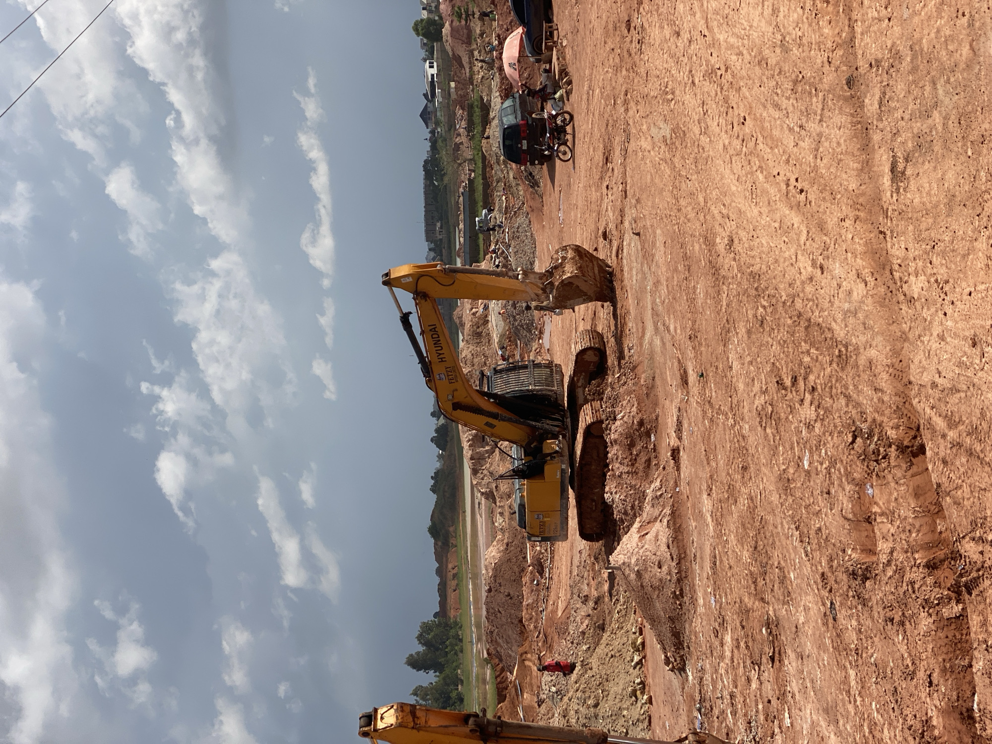 The image depicts a construction site in Reifild-Ist, Nigeria. A large yellow excavator is the primary object in the foreground, its arm extended as it works on the red earth. The scene is characterized by mounds of soil and a partially dug-out area. In the background, there are several buildings, some appearing to be under construction and others already built. A dark van and a motorcycle are visible near a cluster of people, suggesting ongoing work and activity. The sky is partly cloudy, indicating daytime. The overall impression is of an active construction project, likely for residential or commercial development, in a rural or semi-urban setting. There are no discernible emotional cues or specific time of day indicators beyond it being daylight. Text is visible on the excavator, reading "HYUNDAI".