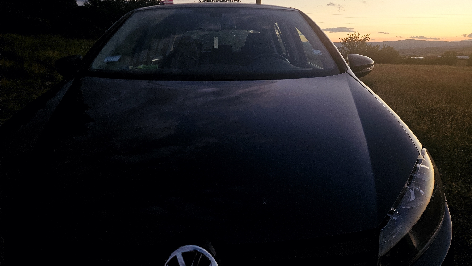 A dark-colored Volkswagen is parked on a grassy field at dusk. The car's hood is the primary subject of the image, taking up most of the foreground. Through the windshield, the interior of the car is visible, including the dashboard, steering wheel, and seats. The background shows a landscape with rolling hills and a warm sunset sky with scattered clouds. The overall lighting suggests either early morning or late evening. The location is listed as Unknown, Unknown.