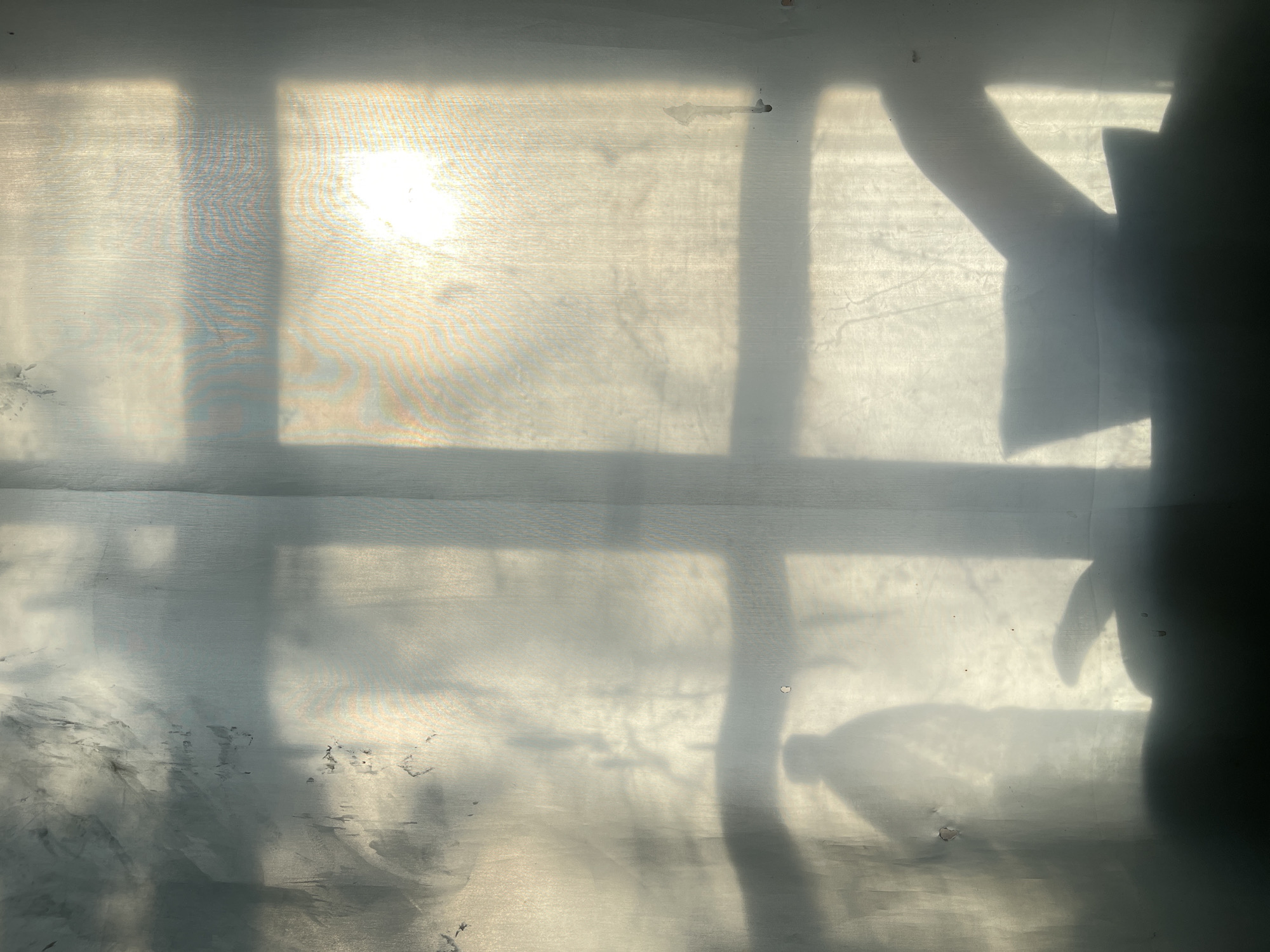 This is a photograph of a semi-transparent white sheet or fabric, likely a curtain or a thin wall, illuminated by sunlight from behind. The sunlight creates a bright, diffused glow, highlighting the texture of the fabric and casting shadows.
The subjects visible are primarily shadows. On the right side of the image, a dark shadow of a person's head and shoulder is prominent, with a hint of an arm reaching upwards. To the left of this, a fainter, elongated shadow appears to be that of a plastic bottle. In the background, the sunlight itself is visible, appearing as a bright, blown-out white area, possibly indicating the sun or a very strong light source. There are also distinct shadow lines crossing the fabric, suggesting the presence of window frames or blinds.
The overall scene appears to be indoors, with the light source coming from outside. The texture of the fabric shows some imperfections, including small dark marks and creases, which are visible through the translucent material. The play of light and shadow creates an abstract and somewhat moody atmosphere. The time of day is likely daytime, given the strong sunlight. There are no visible text elements in the image. The mood evoked is one of subdued light and shadow, with a sense of domestic interiority.