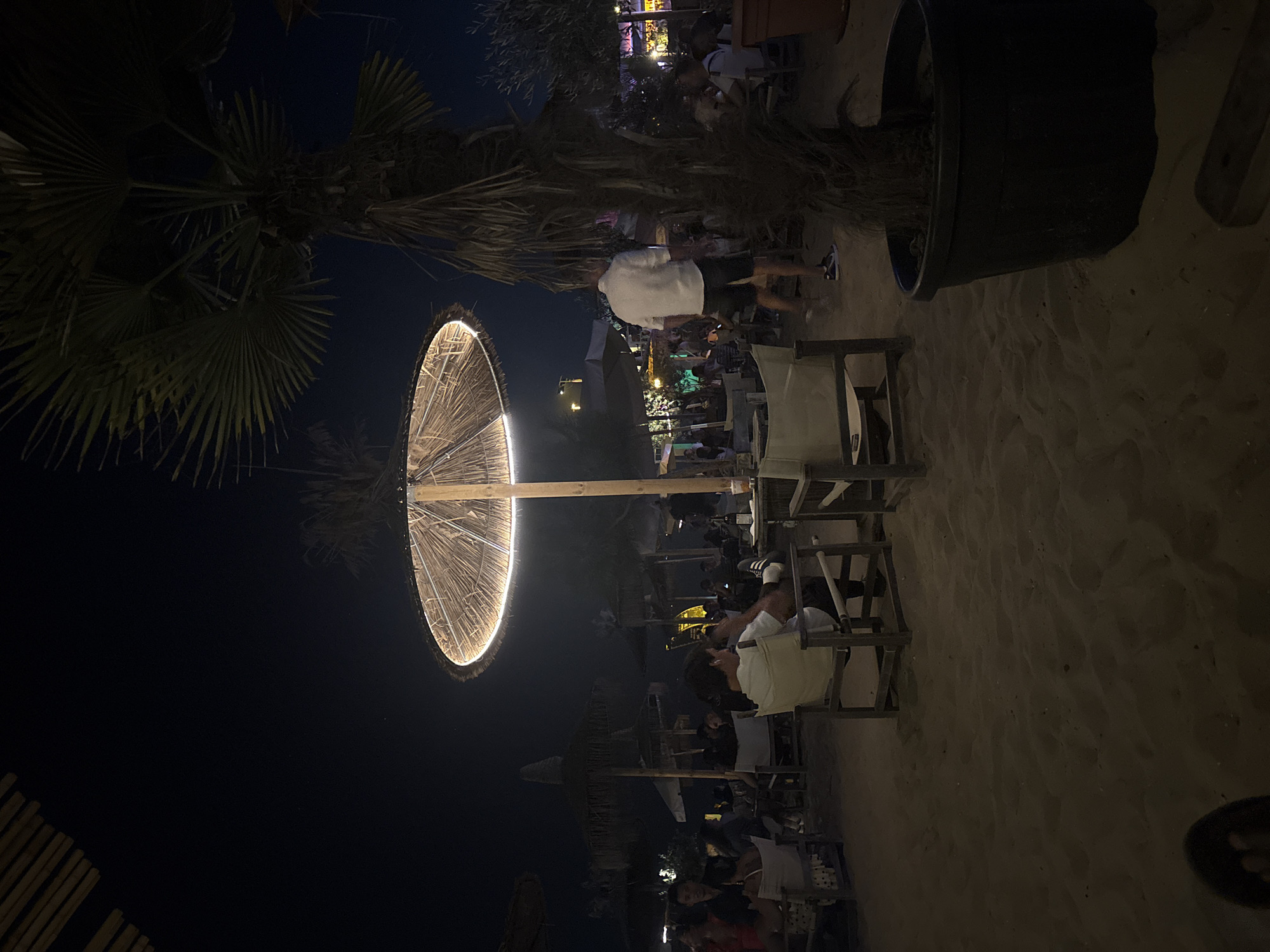 The image depicts a beachside lounge scene at night. In the foreground, the sandy ground is visible, with several chairs and tables arranged for patrons. A large, straw-like umbrella with integrated lighting illuminates the central area. A palm tree stands to the right of the umbrella, its fronds casting shadows. In the mid-ground, people are seated at tables and mingling, suggesting a social gathering or dining experience. The background is dark, indicating nighttime, with some ambient lighting from distant structures and possibly more umbrellas. The overall atmosphere appears relaxed and recreational, typical of a beach bar or restaurant.