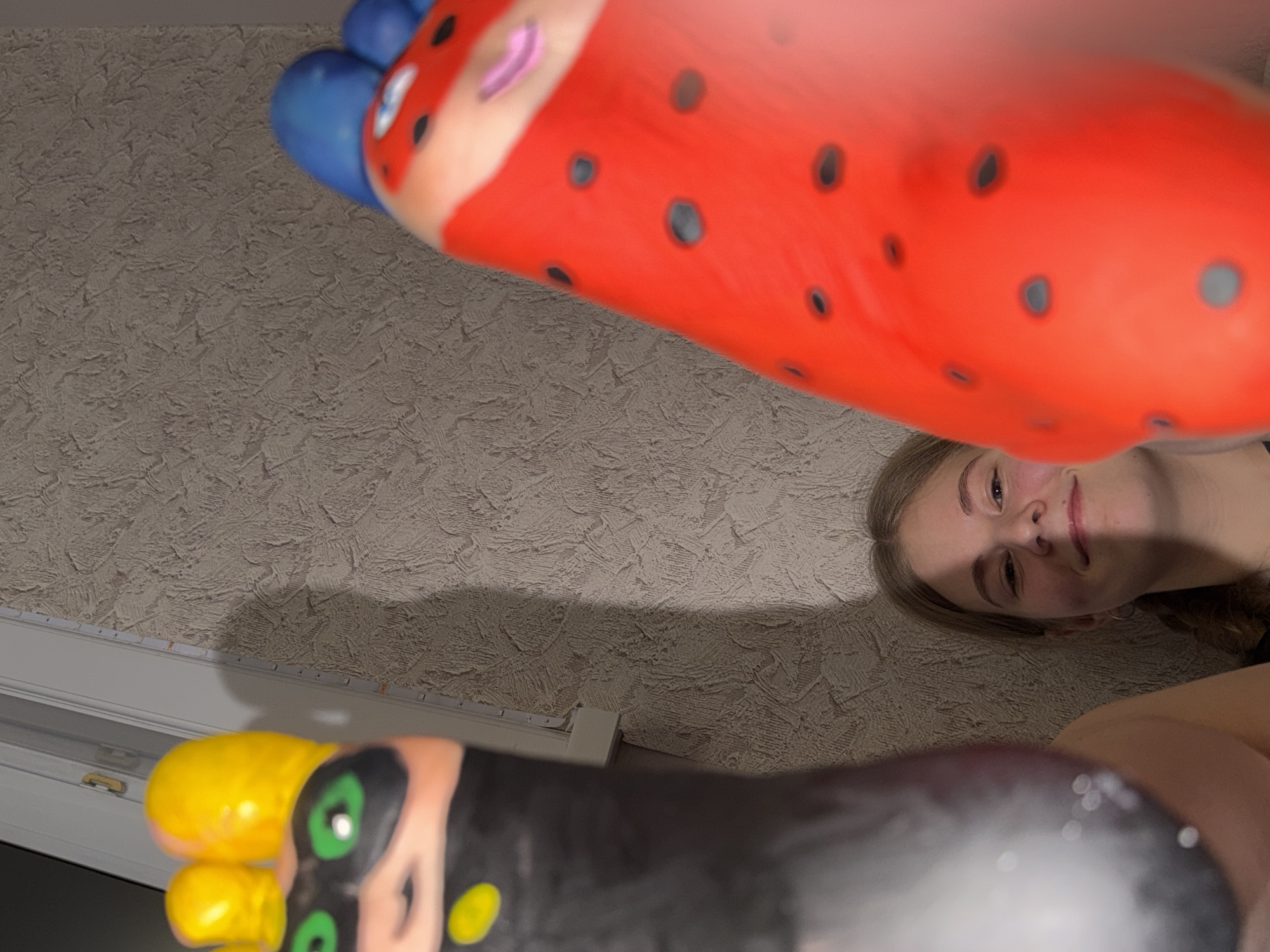 A low-angle shot captures a woman with fair skin and light brown hair looking up and slightly to the right. She appears to be lying down. In the foreground, two brightly colored, possibly inflatable, objects obscure the view. The object closer to the top of the frame is predominantly red with black polka dots and has blue sections. The object closer to the bottom of the frame is black with yellow and green sections. The background consists of a textured, light-colored wall and a white window frame. The location is Perm, Russia.