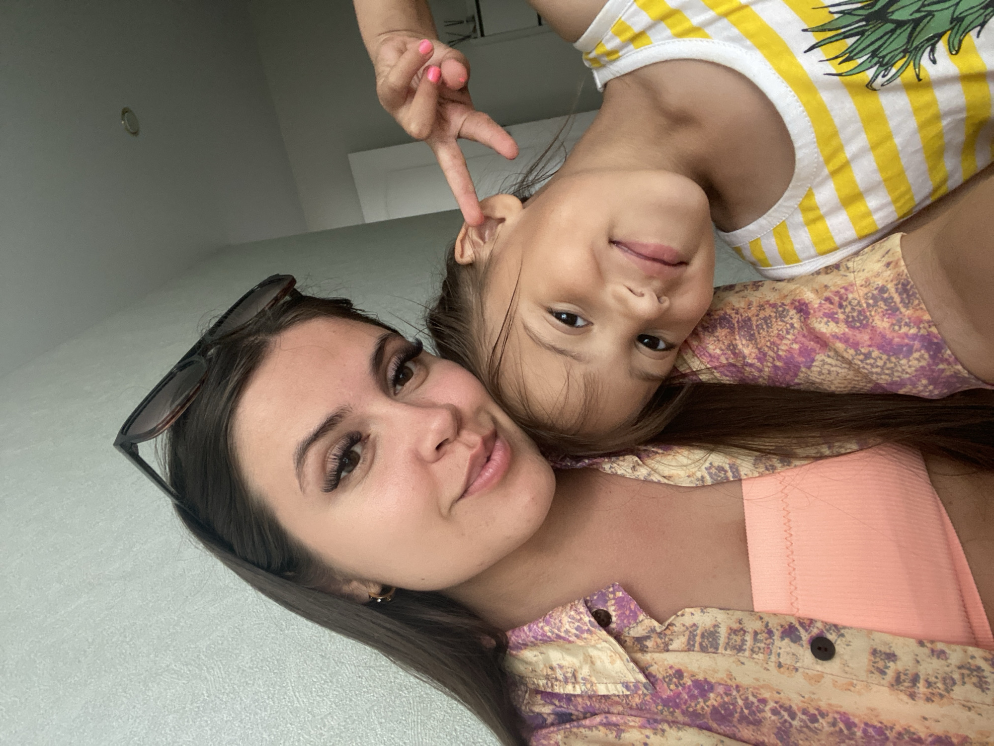 In this close-up selfie, two females, an adult and a child, are positioned in the foreground. The adult, on the left, has long, dark hair, makeup, and sunglasses pushed up on her head. She is wearing a patterned shirt over an orange tube top. The child, on the right, has dark hair and is wearing a yellow and white striped tank top with a pineapple graphic. The child is making a peace sign with her fingers near her ear and leaning her head on the adult's shoulder. They are indoors, with a lightly textured wall and part of a doorway visible in the background. The lighting suggests an indoor setting, possibly a home. There is no indication of the city of Chaikovsky, Russia, or any specific location details.