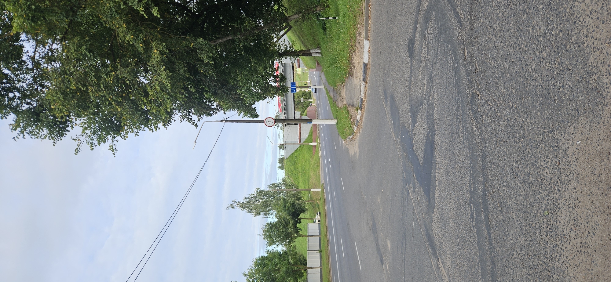 The image shows a street intersection in Vitebsk, Belarus. A road with multiple lanes runs horizontally across the frame, with a pedestrian walkway on the right. Green hills rise on either side of the road, topped with trees and a bridge. A speed limit sign with the number "41" is visible on a lamppost. The sky is overcast. The road surface appears worn, with patches of darker asphalt and some visible cracks. Trees with green foliage line the sides of the road and the hills. A white car is parked on the left side of the road. The scene appears to be a typical urban streetscape on an overcast day.