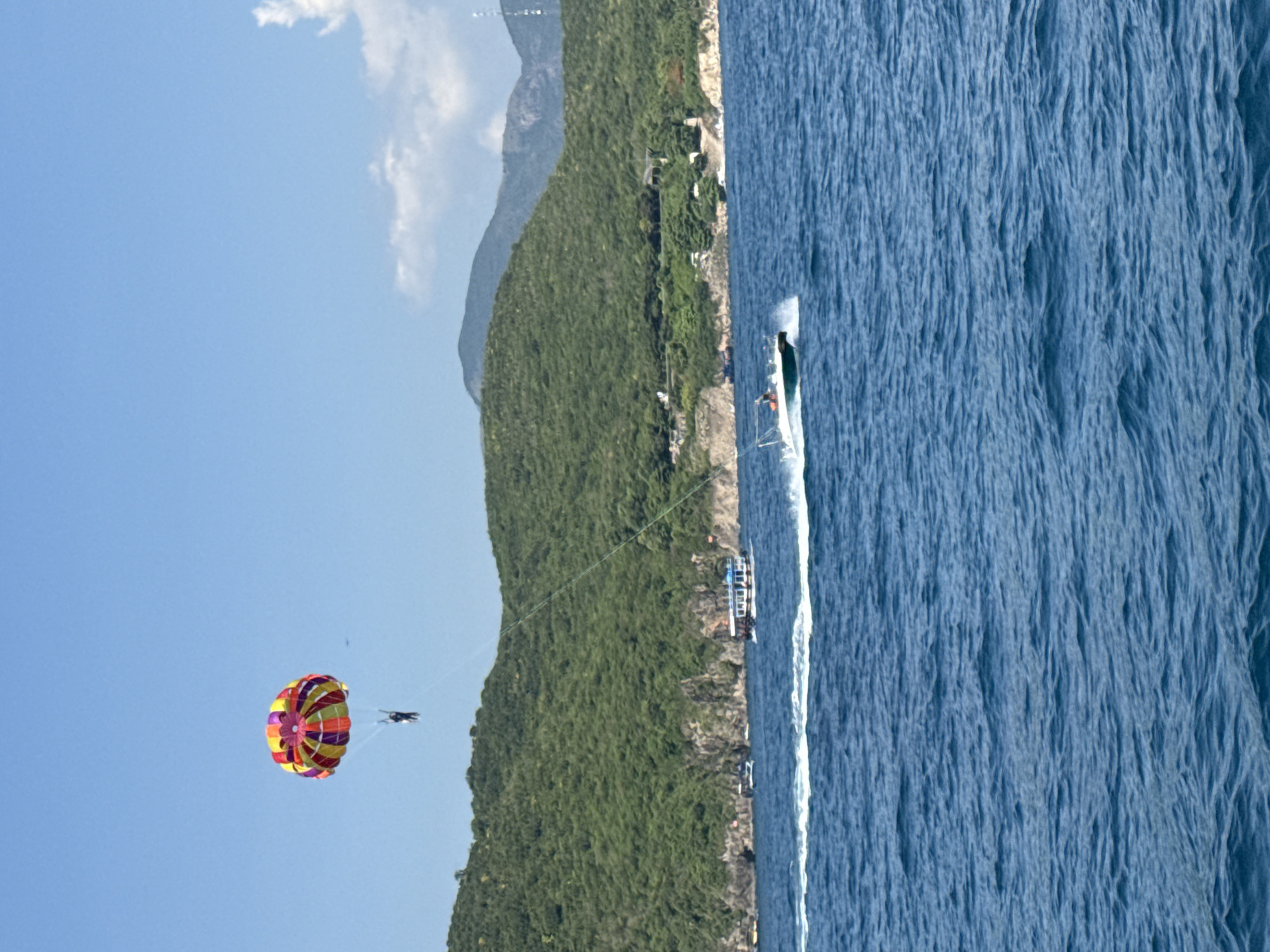 The image depicts a parasailing activity off the coast of Bac Nha Trang Ward, Vietnam. A person is suspended in the air by a brightly colored parasail, which is pulled by a boat in the water below. The boat is creating a white wake as it moves across the blue ocean. In the background, a lush green, tree-covered island rises from the sea, with some rocky cliffs visible along its shore. A few other boats are anchored near the island. The sky is a clear, bright blue with a few scattered white clouds. The overall scene is one of a sunny day at a tropical beach resort.
