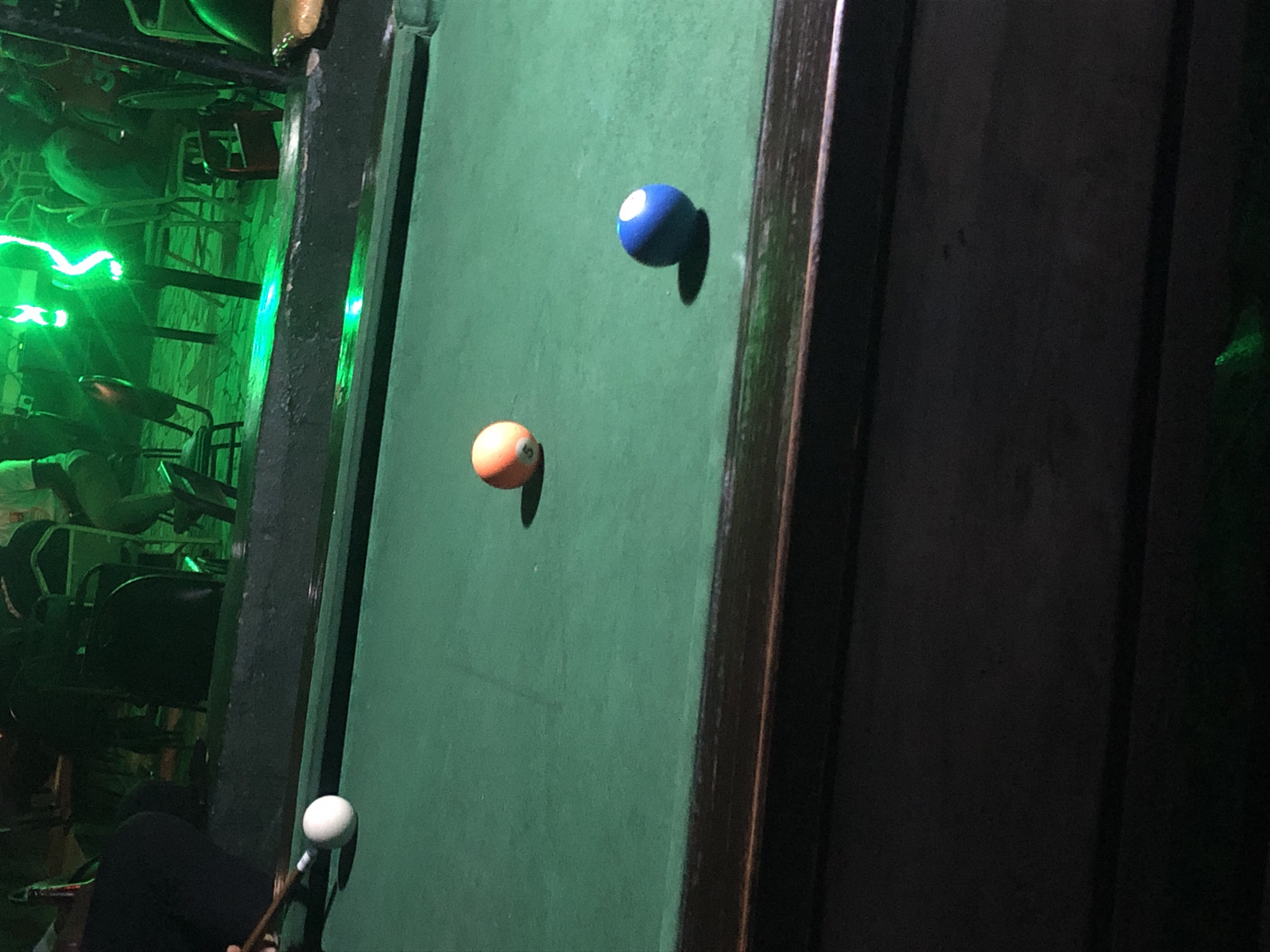 A billiard table is shown in a dimly lit establishment with green neon lighting in the background. Three billiard balls are visible on the green felt surface. A white cue ball is on the left side of the frame, with a pool cue aimed at it. Further to the right, an orange ball with the number "5" is present. A blue ball with a white stripe is to the right of the orange ball. The table is bordered by a dark wooden edge. In the background, shadowy figures and chairs can be seen, illuminated by the green neon light.
