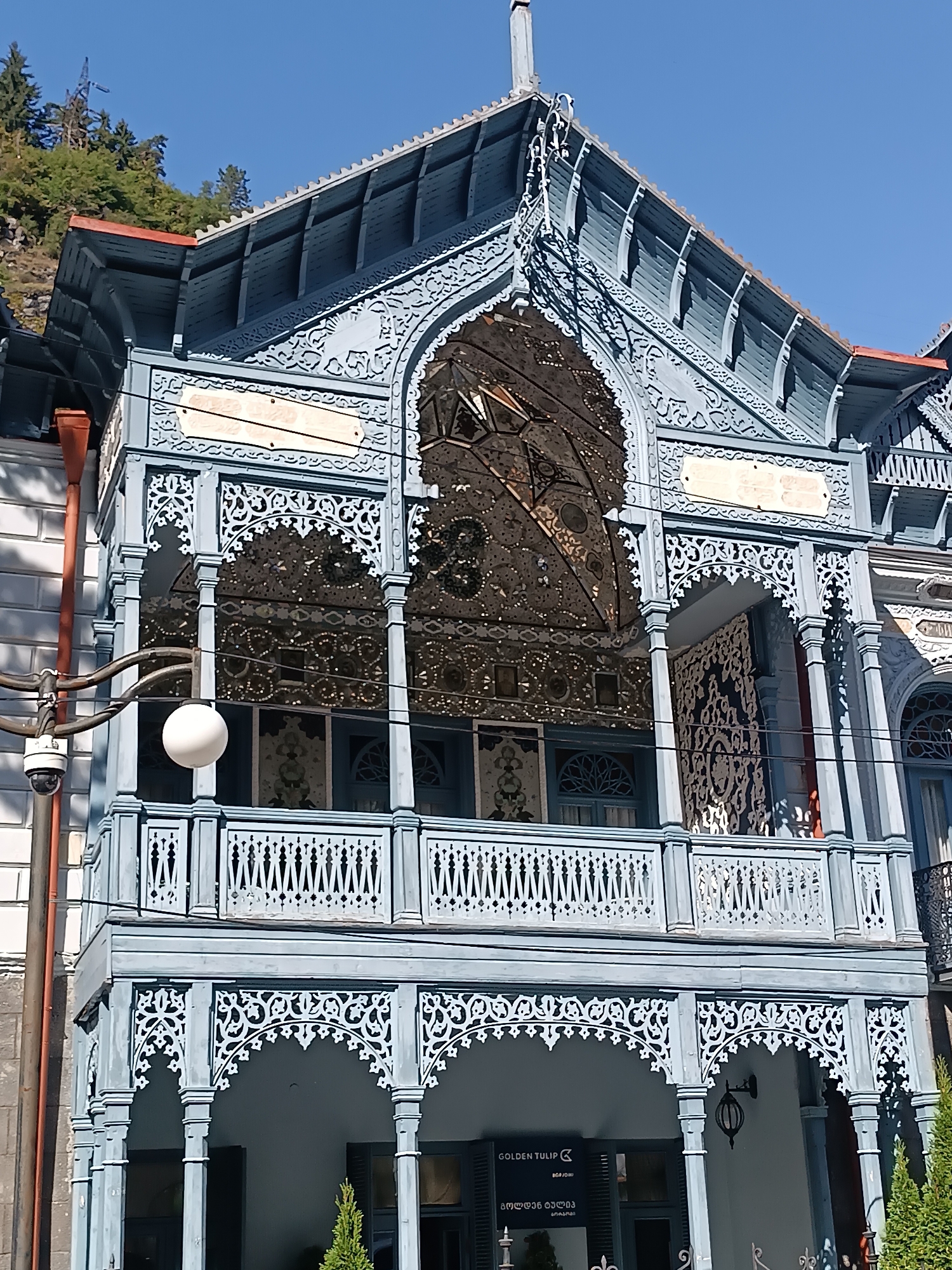 The image shows a close-up, eye-level shot of the ornate wooden balcony of a building, likely in Borjomi, Georgia. The balcony is painted a light blue and features intricate latticework and decorative carvings. The roof above the balcony is also blue, with a red trim on the eaves. Behind the balcony, a white building with a textured facade is visible. A street lamp with a white spherical shade hangs from a dark metal pole attached to the side of the building, and a security camera is mounted on the same pole. The sky is clear and blue. Below the main balcony, another level of the building features arched openings with similar decorative wooden elements. A sign is visible below the arches, partially obscured by foliage, that reads "GOLDEN TULIP" and some Georgian text. A small evergreen tree stands to the right of the entrance. The overall impression is of a well-preserved, historic building with detailed architectural features.