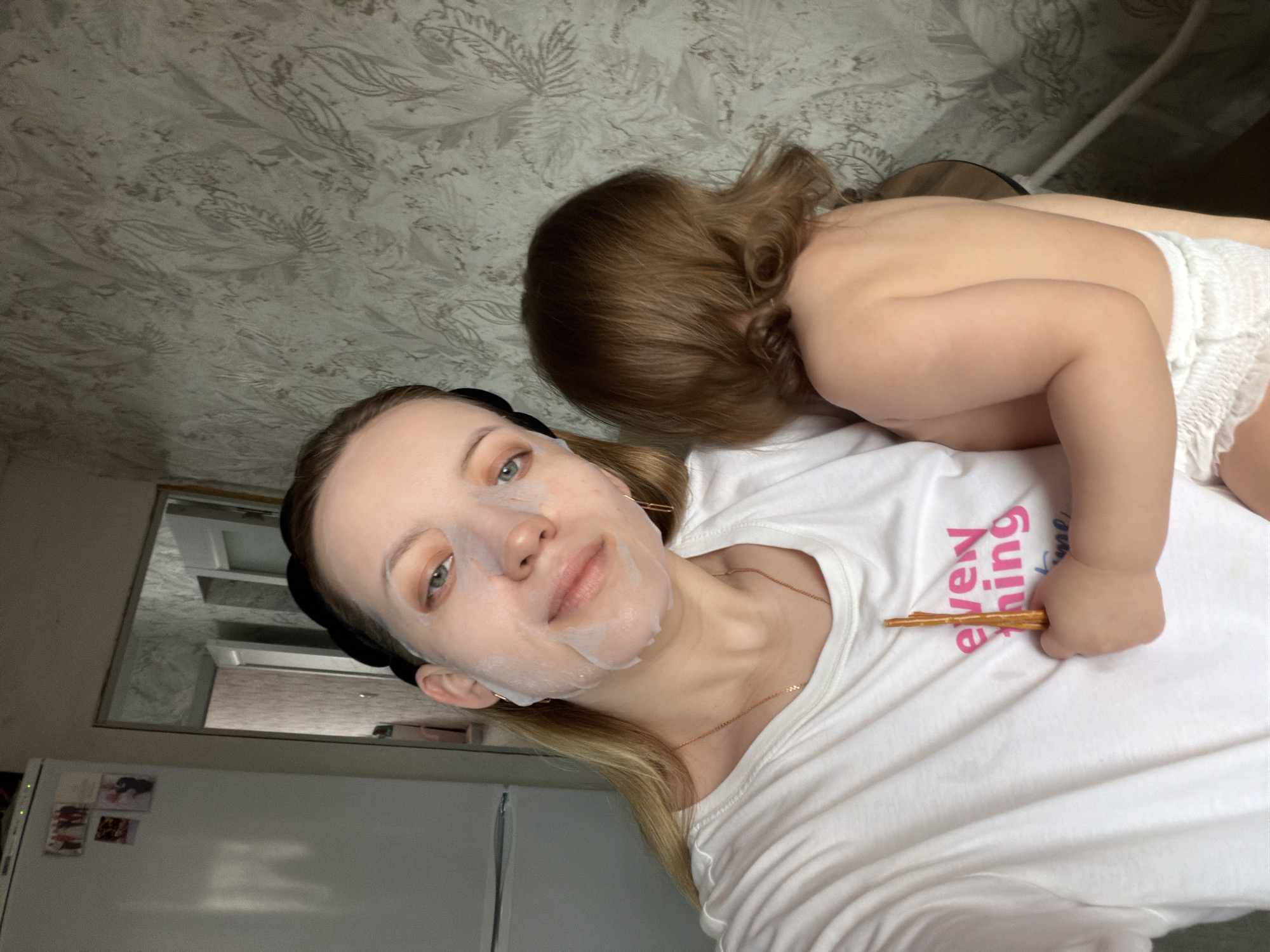 A selfie-style, slightly angled photograph shows a young woman wearing a facial mask and a white t-shirt with the word "everything" in pink lettering, lying down with a toddler resting on her. The child is facing away from the camera, wearing a diaper, with brown curly hair. A stack of pretzel sticks are on the woman's shirt and the child has their hand on it. A white refrigerator is visible in the background, with photographs on the door. The background also shows a wall with a subtle pattern. The location is likely indoors, potentially a home, but it is not clear if it is in Reshetnikovo, Russia.