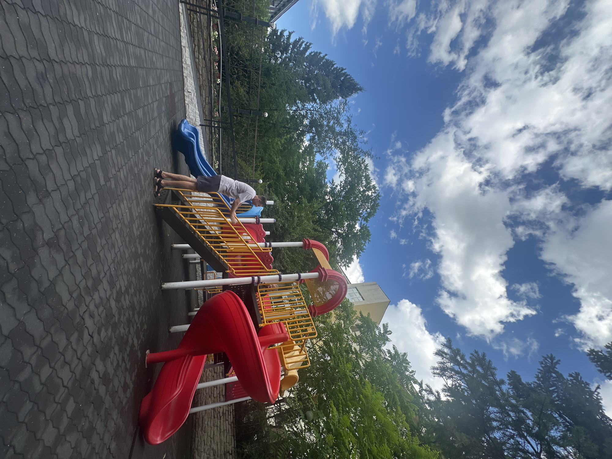 A man stands on a playground in Belokurikha, Russia, facing a brightly colored plastic slide structure. He is wearing a light-colored t-shirt and dark shorts. The playground features a red spiral slide and a blue straight slide, both made of plastic. The ground is covered with black interlocking rubber tiles. In the background, a black metal fence with decorative elements separates the playground from a stone wall and a lush green area with trees. Beyond the trees, a light-colored building with windows is visible. The sky above is a vibrant blue with scattered white clouds. The overall impression is a sunny day at a children's play area.