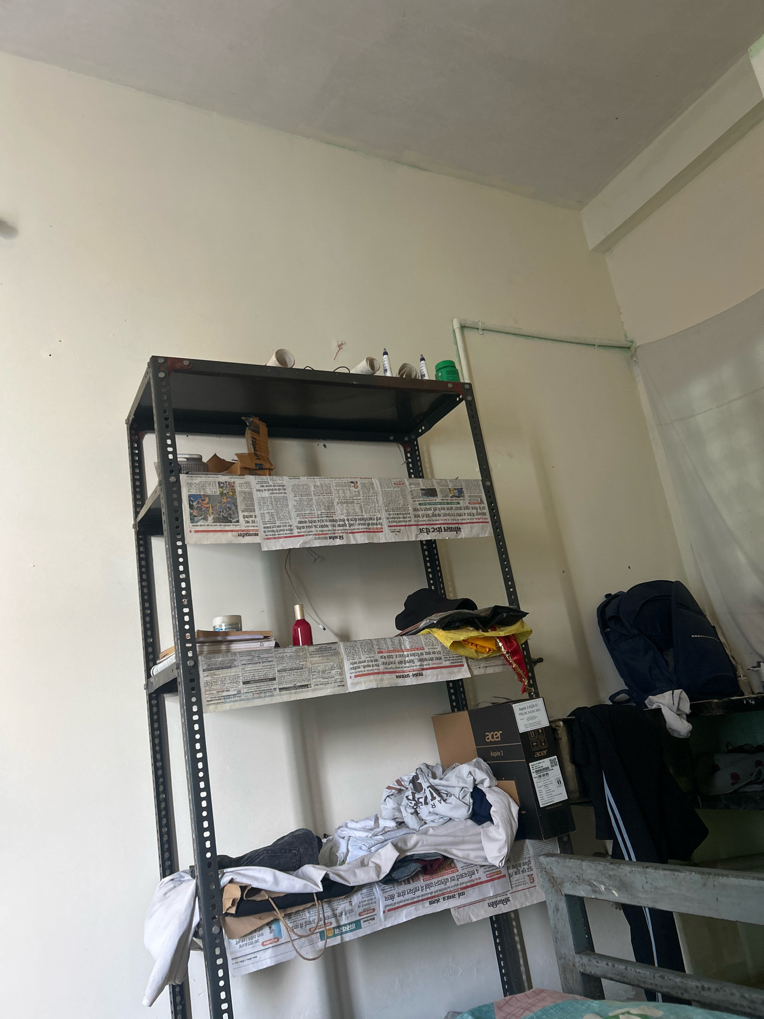 The image shows a cluttered room interior in Bhopal, India. The focus is on a tall, metal shelf unit filled with various items. On the top shelf, there are some paper rolls and pens. The second shelf contains newspapers and some scattered items. The third shelf also holds newspapers, a red bottle, and a roll of tape. On the fourth shelf, there are more newspapers, clothing items, and a cardboard box with the Acer logo. A dark blue backpack is visible on a lower shelf, along with other clothing. The overall environment appears unkempt, suggesting a residential or storage space.