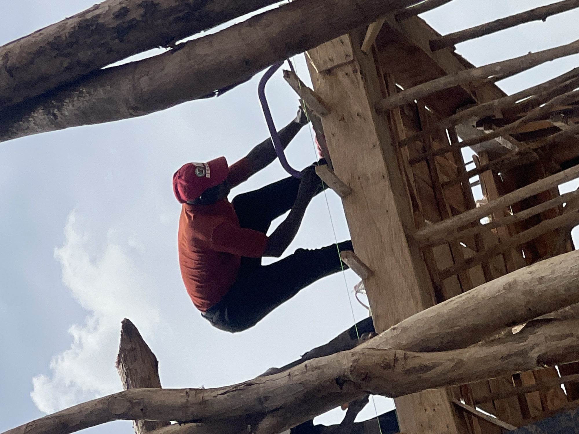 A man in a red cap and orange shirt is climbing a wooden structure in Jos, Nigeria. He uses a purple rope as an aid. The structure appears to be under construction, with roughly hewn wooden beams and posts. The sky is visible through gaps in the roof.