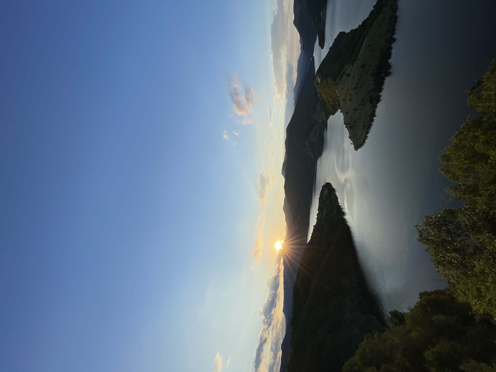 The image depicts a panoramic view of the Borovitsa Dam in Bulgaria at sunset. The sun is low on the horizon, casting a golden glow and radiant beams across the sky and water. The dam's vast reservoir is visible, reflecting the warm colors of the sunset. Surrounding the water are rolling, tree-covered hills that gradually recede into the distance, forming layers of mountains against the blue sky. The sky is clear with a few wispy clouds tinged with pink and orange from the setting sun. In the foreground, the lush green foliage of trees frames the lower portion of the image, adding depth and a sense of being at an elevated viewpoint. The scene is serene and natural, with no people or artificial structures prominently featured.