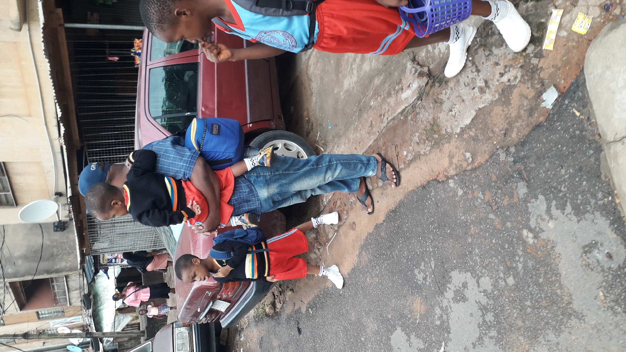 In a daytime street scene in Onitsha, Nigeria, a man wearing a blue baseball cap, a blue checkered shirt, and blue jeans carries a young child wearing a red and black outfit. Two other young children in similar red and blue school uniforms stand nearby. A burgundy sedan is parked beside them. The street appears to be paved and is partially wet. Buildings with corrugated metal roofs and satellite dishes are visible in the background. Some people are standing in front of one of the buildings. One of the children carries a blue plastic basket.