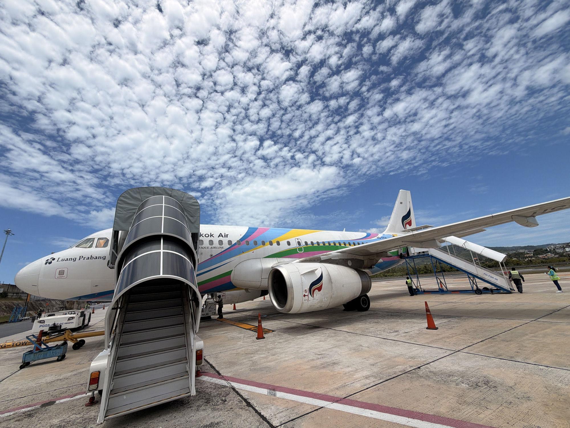 An airplane with the "Bangkok Air" livery and "Luang Prabang" text on its fuselage is parked on an airport tarmac under a bright blue sky filled with scattered white clouds. A mobile boarding staircase is positioned at the front of the aircraft, and another staircase is connected to the rear exit. Several airport workers, some wearing high-visibility vests, are visible near the aircraft. In the background, there are buildings, trees, and distant hills. The tarmac is marked with painted lines.