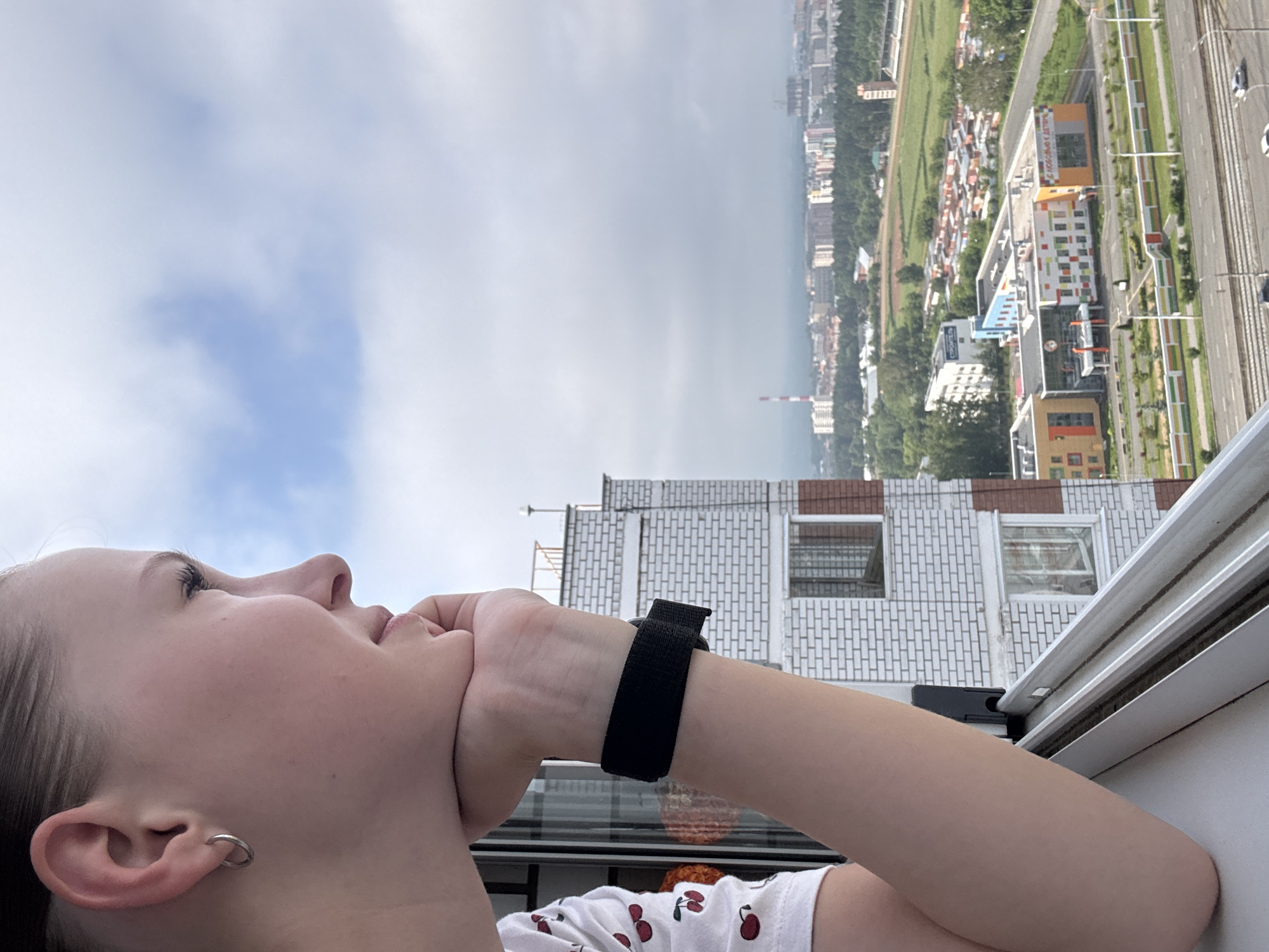 A young woman is captured from the side, resting her chin on her hand and looking out a window. She has a silver hoop earring in her left ear and is wearing a white t-shirt with a cherry pattern and a black wristband. Her arm is resting on the window frame. Outside, a cityscape unfolds with apartment buildings, some with white brick facades, and a distant view of industrial structures and green spaces. The scene is set on a cloudy day, with the sky appearing overcast. The location is confirmed to be in Izhevsk, Russia, specifically from a high vantage point, likely an apartment building. The image is a portrait of a contemplative moment against an urban backdrop.