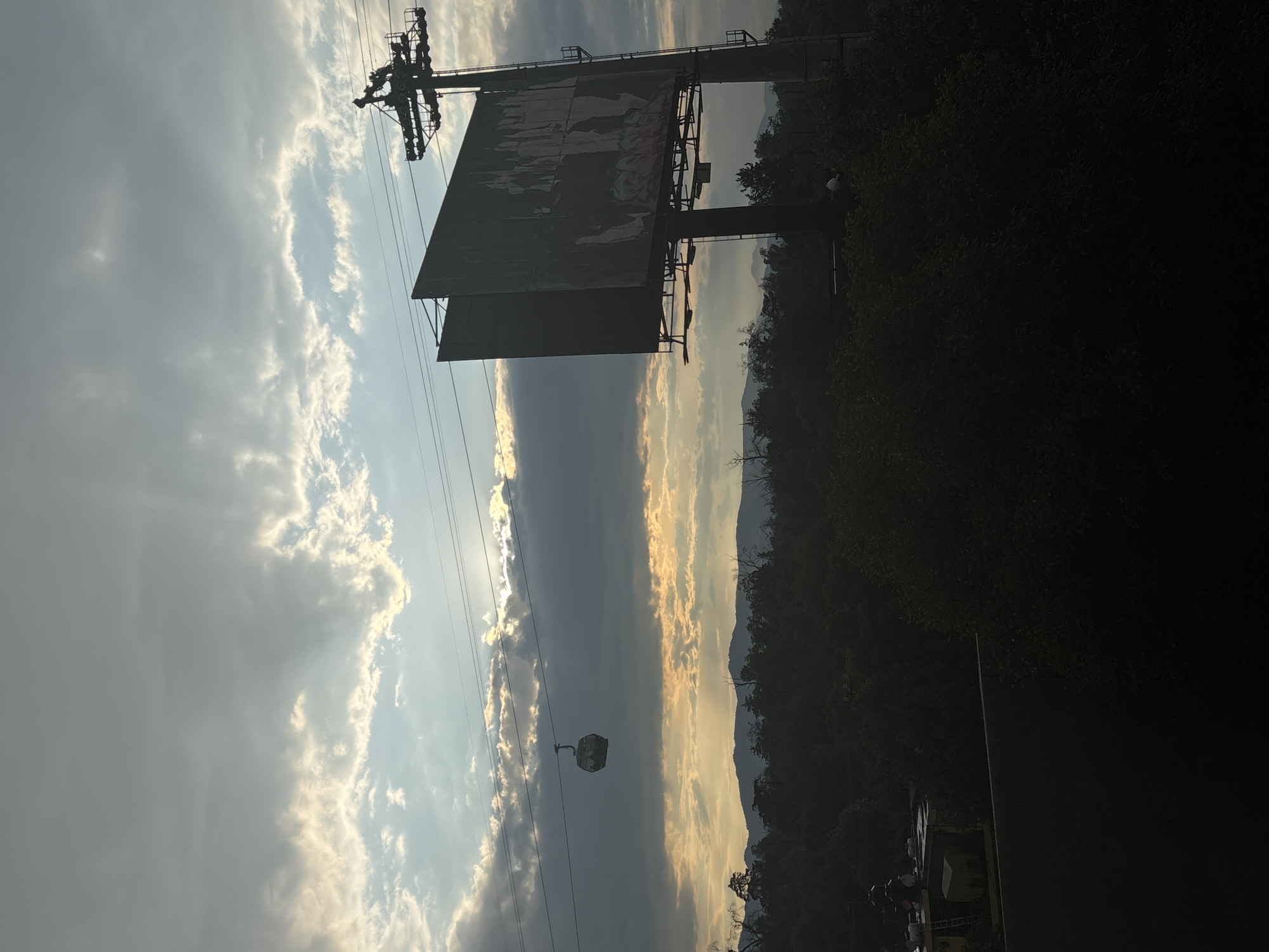 The image shows a landscape in Mexico City, Mexico. The sky is filled with clouds, some illuminated by the sun, while others are dark. A cable car and its supporting structure are prominent on the left side of the image. A large, weathered billboard stands near trees. In the background, a mountain range is visible under the sunlight. The foreground is mostly dark.
