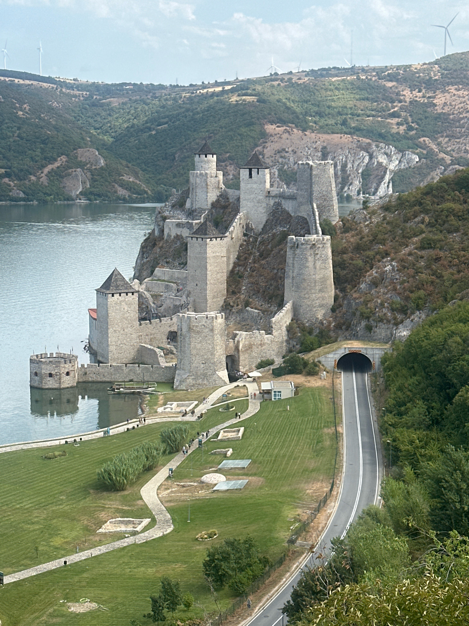 An elevated, full shot captures Golubac Fortress, a medieval castle situated on the Danube River's edge in Golubac Municipality, Serbia. The fortress, built of stone, features numerous towers of varying heights and designs, some with pointed roofs. A prominent round tower stands at the water's edge, seemingly integrated with the river. The castle is perched on a steep, rocky incline, covered with sparse vegetation.
Below the fortress, a well-maintained grassy area with paved pathways and scattered archaeological remnants is visible. Several small groups of people are dispersed throughout this area, walking along the paths or observing the historical site. A road curves from the foreground, passing through a tunnel carved into the hillside to the right of the fortress. The tunnel is illuminated, and its entrance is marked with yellow and black diagonal stripes.
Beyond the fortress, the Danube River extends into the distance, reflecting the overcast sky. The surrounding landscape is characterized by rolling hills covered in dense green forests and patches of dry grass. Wind turbines are visible on the distant hills. The overall scene depicts a historical landmark integrated with modern infrastructure and natural scenery.