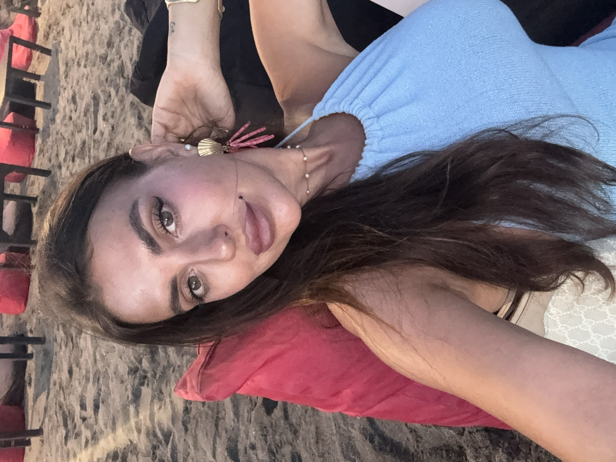 A woman with dark hair and light skin is lying on a red pillow in an outdoor setting, possibly a beachside area. She is wearing a light blue top, jewelry including earrings and a necklace. In the background, there is sand and chair frames with red cushions. Based on the prompt's request for the location, this scene could potentially be within North Kuta, Indonesia, though this cannot be conclusively determined from the image alone.